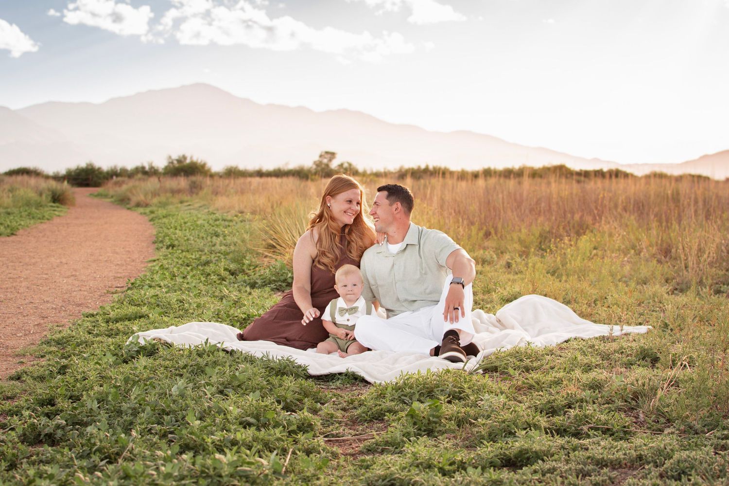 Family sitting on white blanket in green field with mountain backdrop at golden hour.