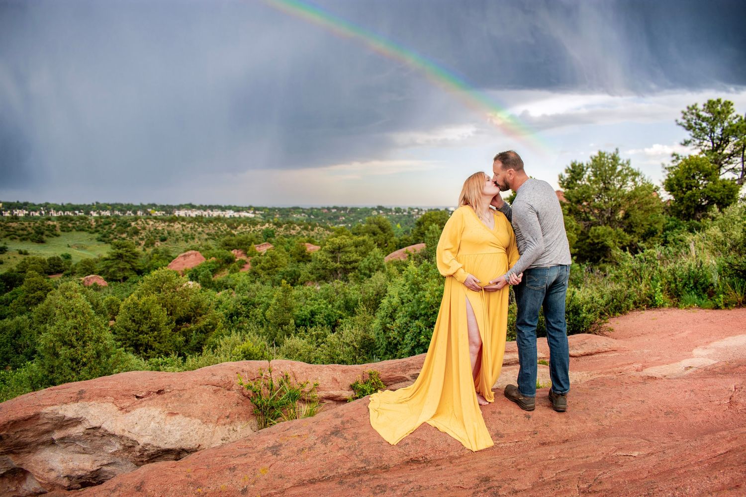 Pregnant couple in yellow gown kiss on red rocks under a rainbow with lush green landscape backdrop.