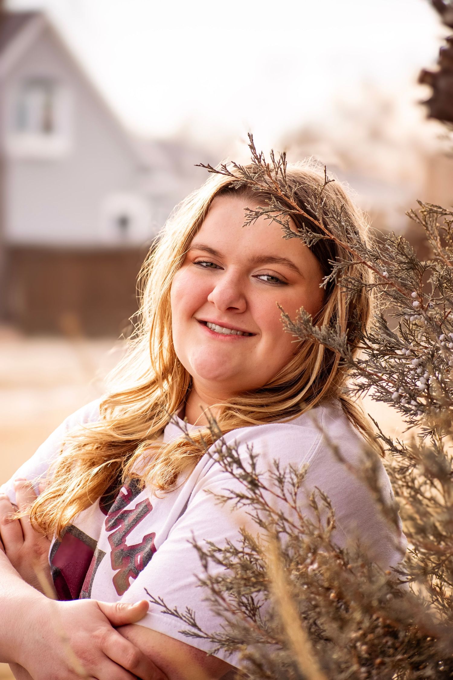 Smiling young woman with blonde hair wearing a natural twig crown, embracing dried plants outdoors near a house.