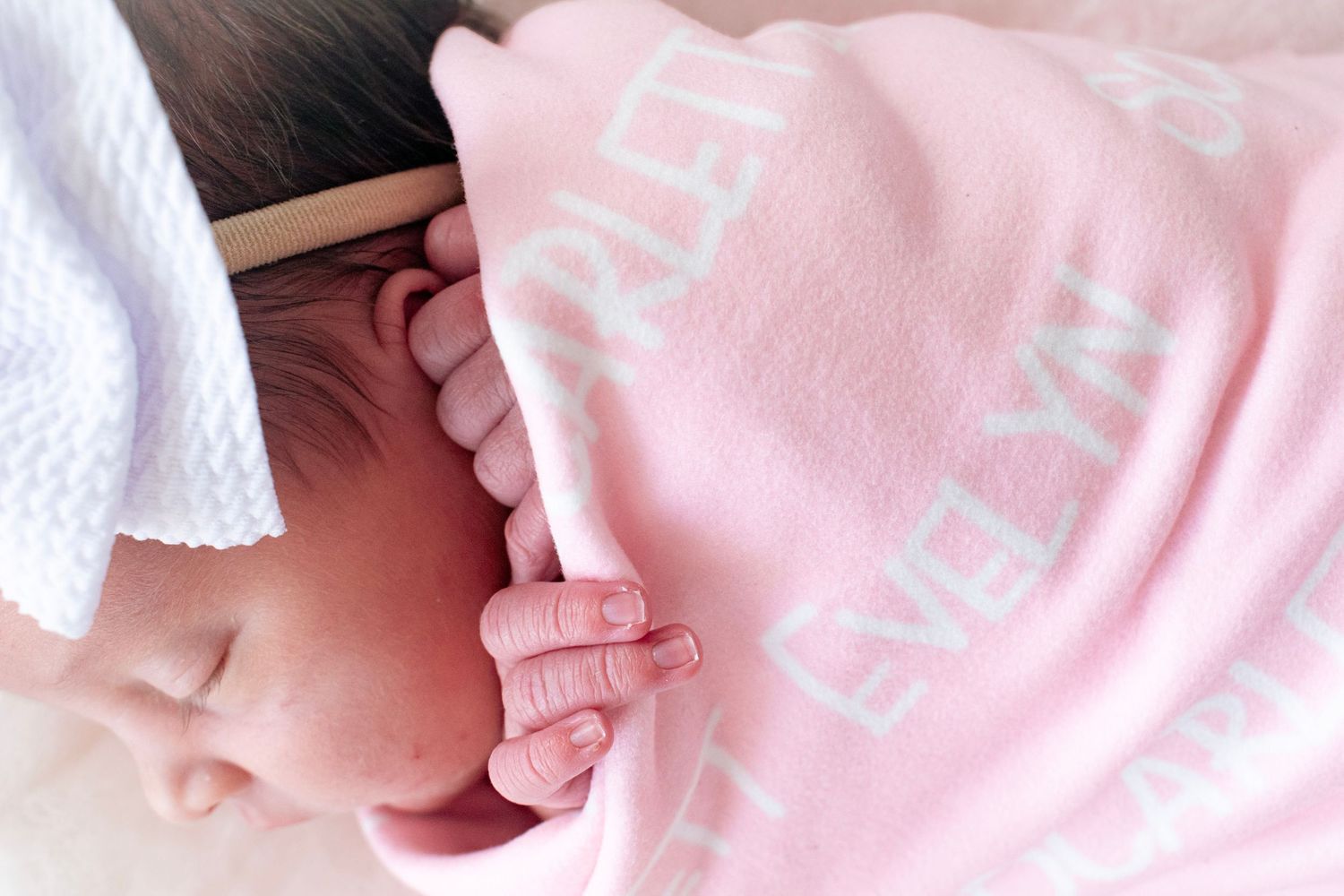 Newborn baby sleeping peacefully wrapped in a personalized pink blanket with white bow headband.