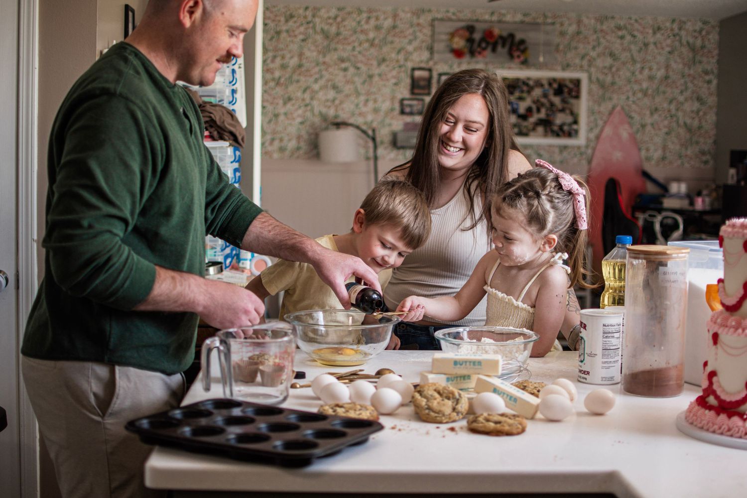 Family baking together in kitchen, parents and two young children mixing ingredients in bowls on white counter.