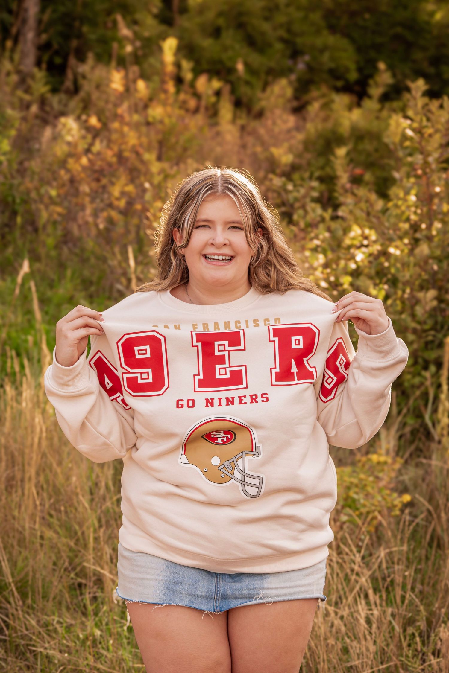 Smiling young woman wearing a San Francisco 49ers cream sweatshirt, posing outdoors in a natural setting.