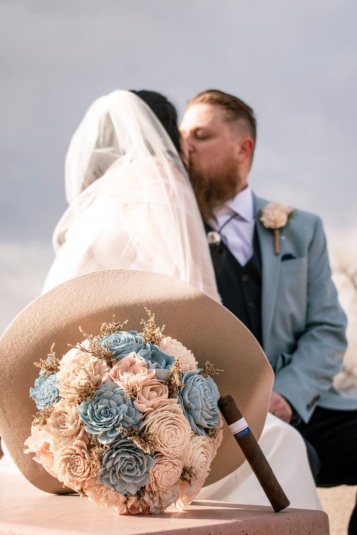Bride and groom kissing behind a bouquet of blue and pink roses with a tan hat at an outdoor wedding.