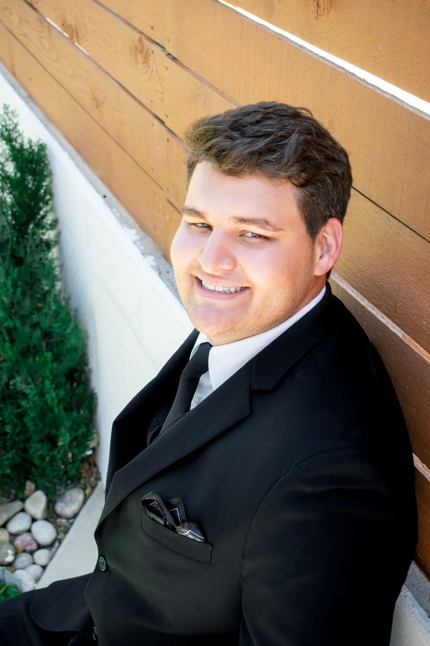 Young man in black tuxedo smiling outdoors, leaning against wooden wall with green shrubs nearby.