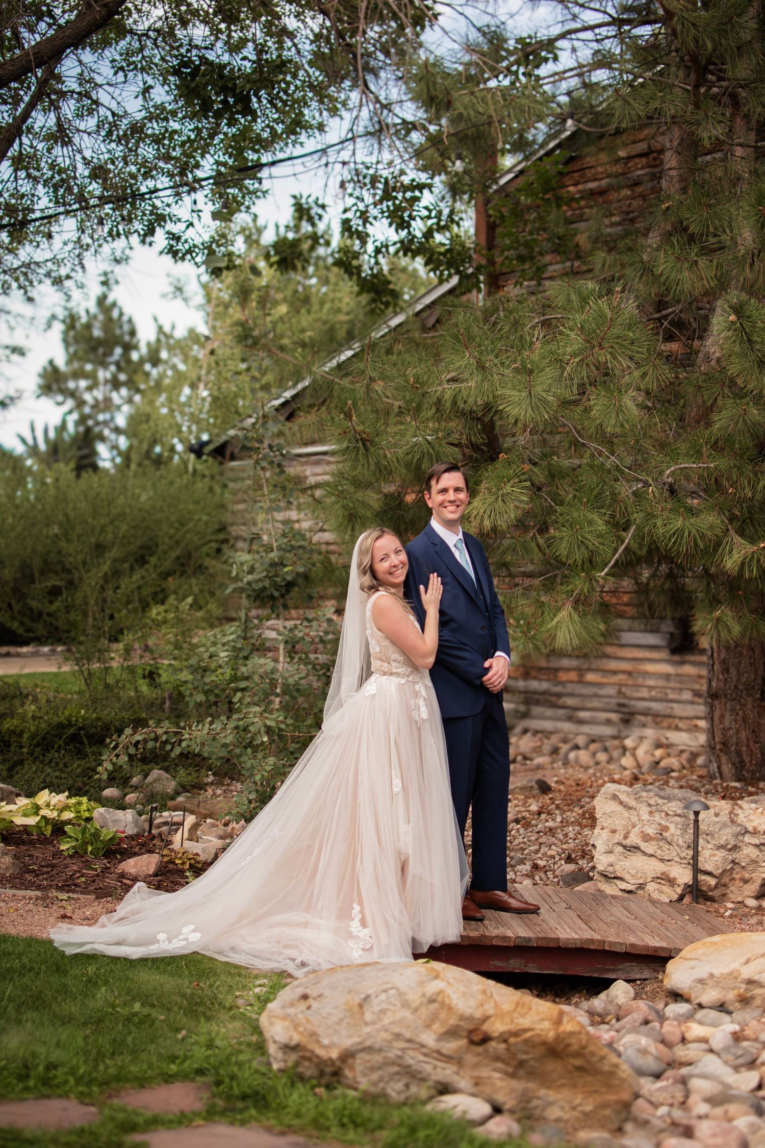 Bride in white gown and groom in navy suit pose on wooden bridge surrounded by lush greenery and stone walls.