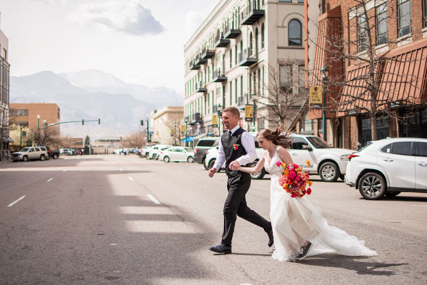 Bride and groom crossing a city street with mountains in background, bride holding vibrant bouquet.