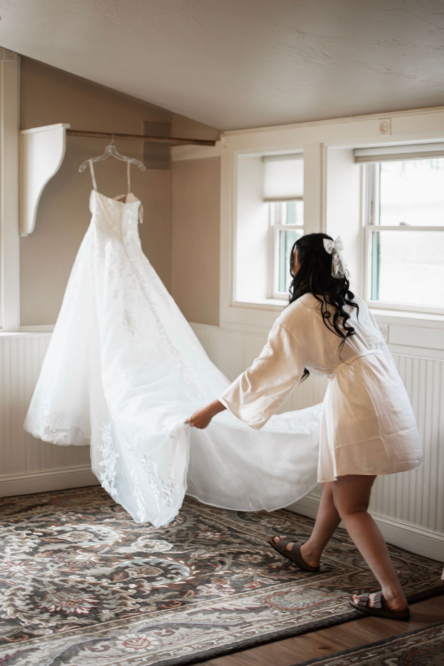 Bride in white robe admiring her lace wedding gown hanging in a sunlit room before the ceremony.