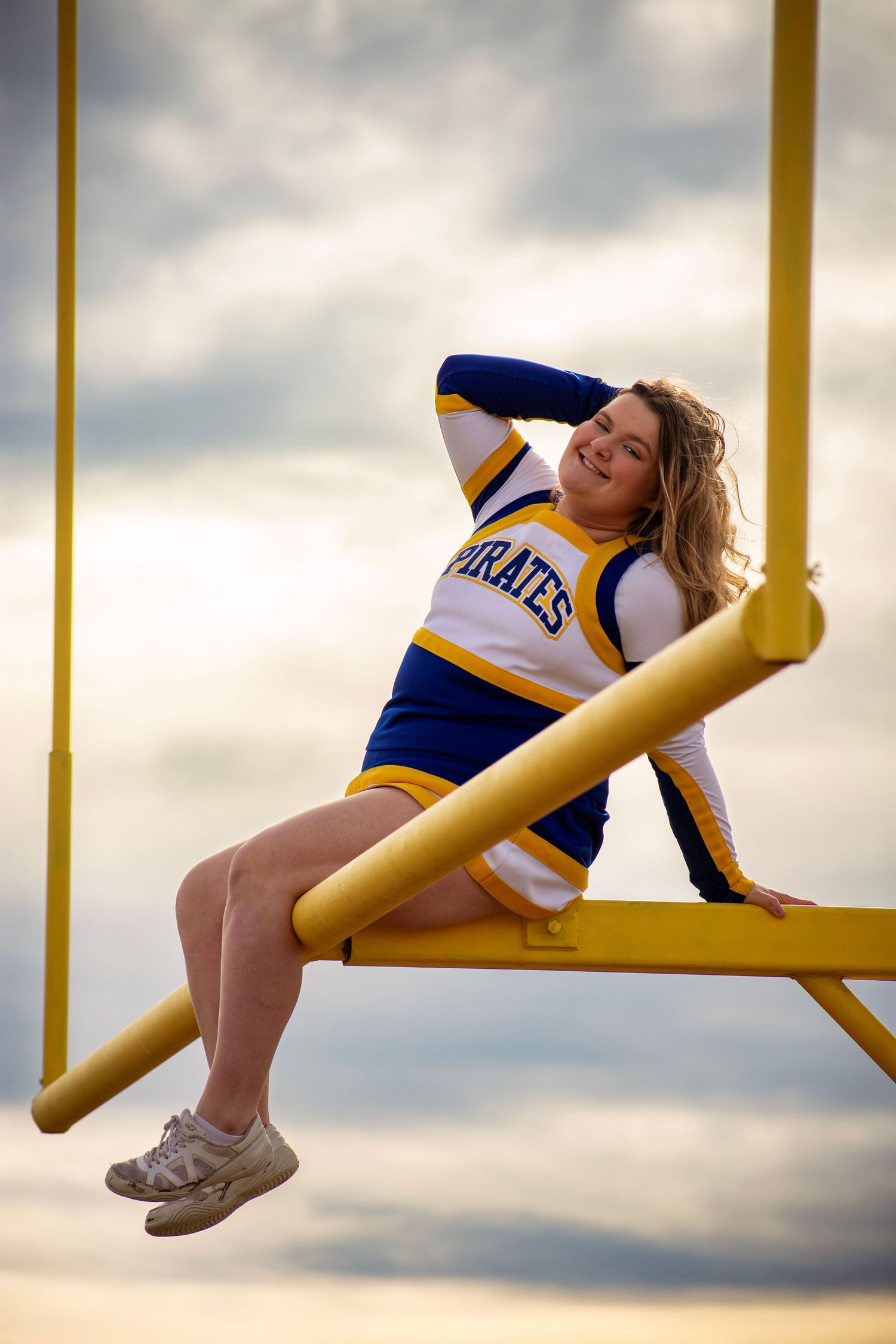 Cheerleader in Pirates uniform sitting on yellow football goal post against cloudy sky.