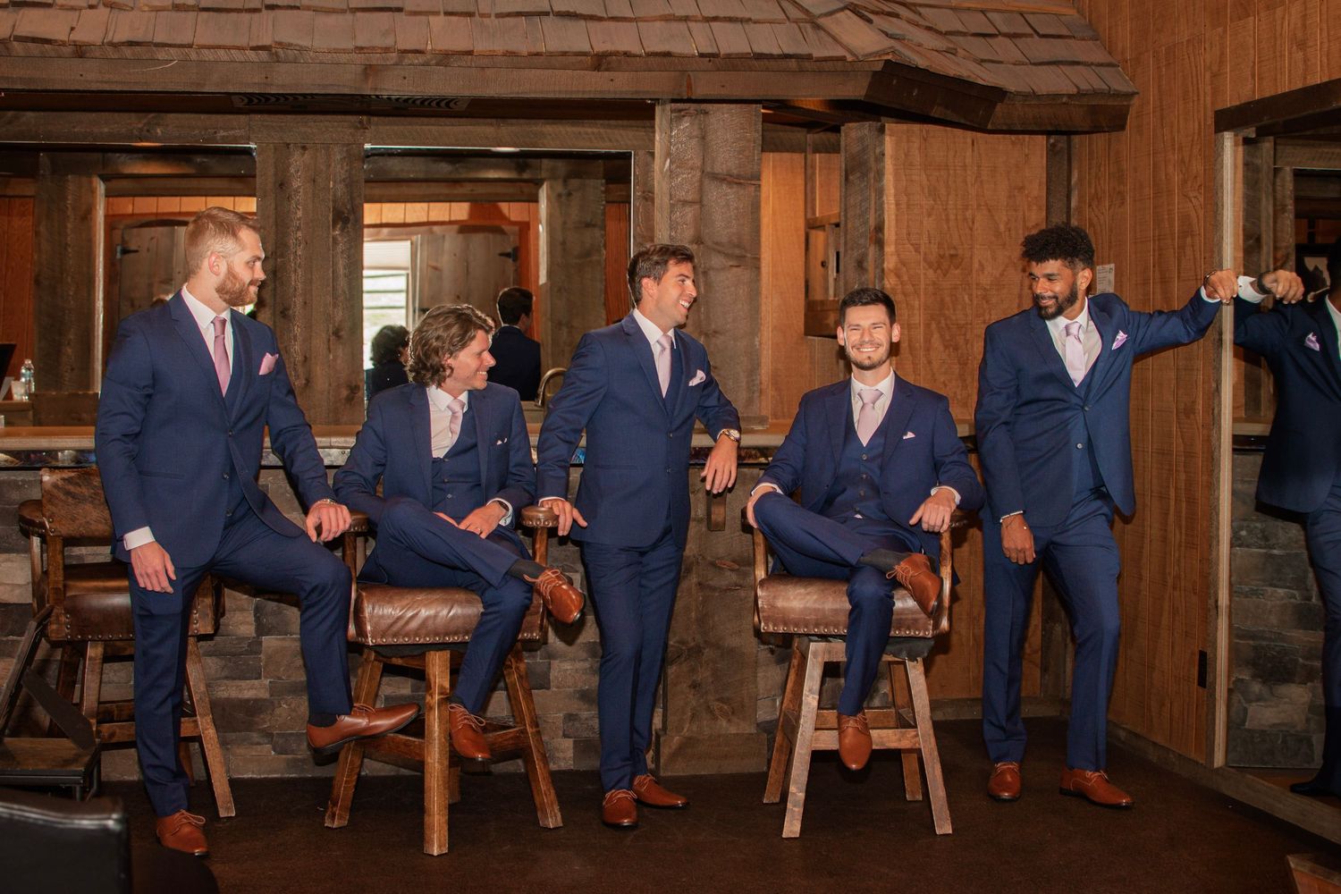 Five groomsmen in navy blue suits pose casually on bar stools in a rustic wood-paneled venue.