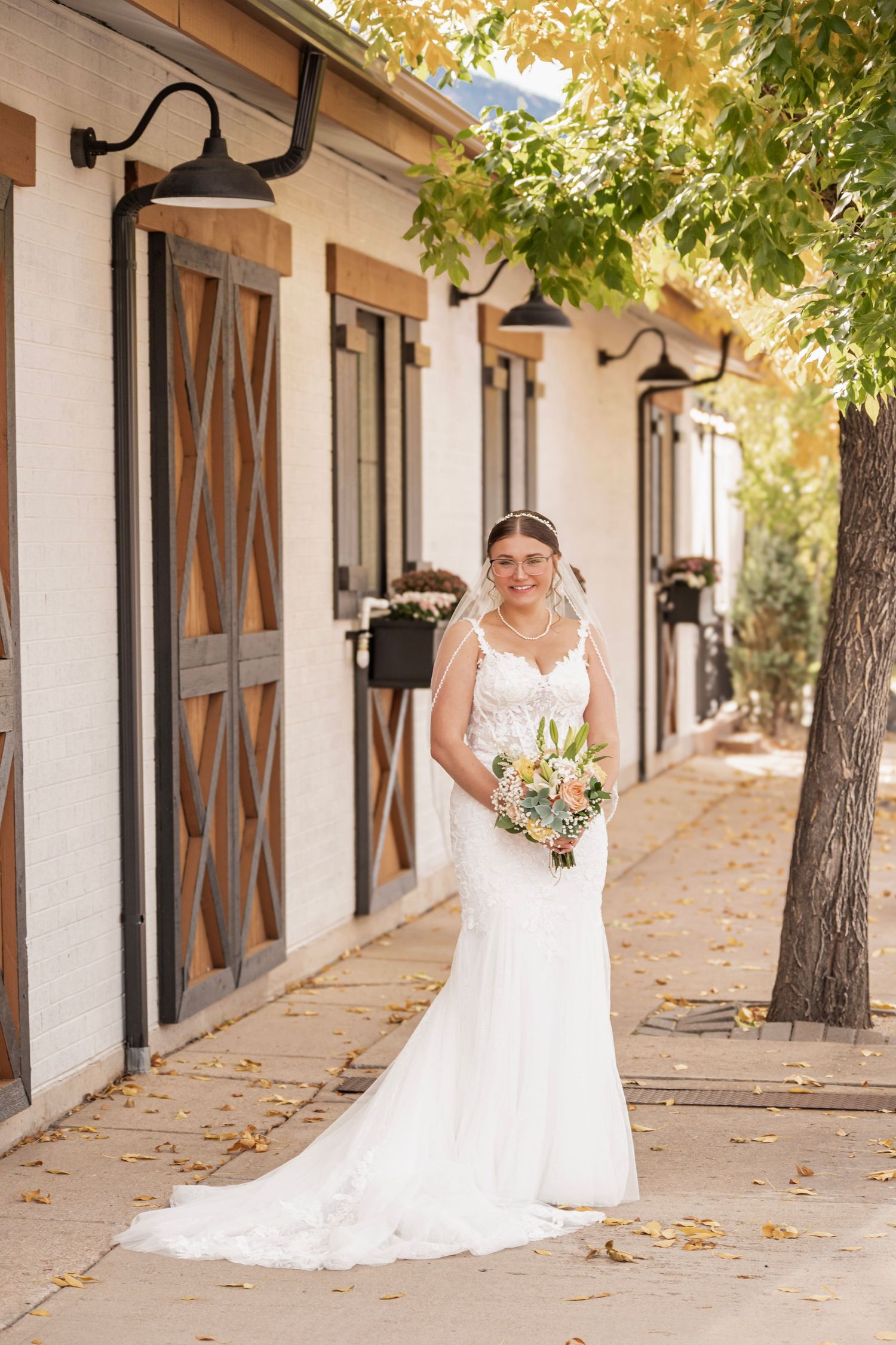 Bride in white lace mermaid gown holding floral bouquet stands on a pathway beside a rustic building with wooden doors.