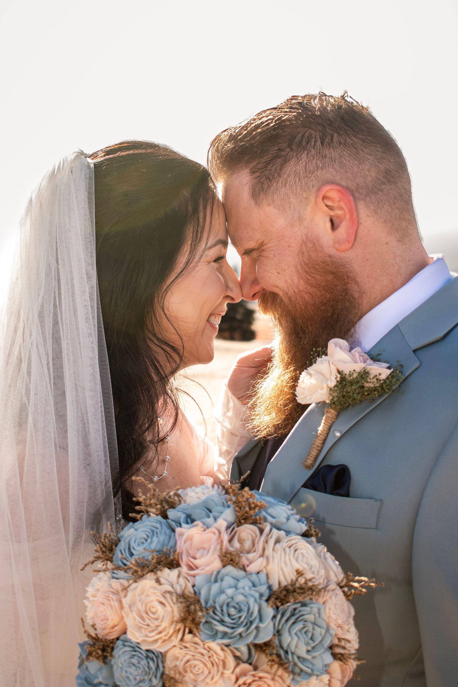 Bride and groom touching foreheads, smiling, with a blue and pink floral bouquet, backlit by golden sunlight.