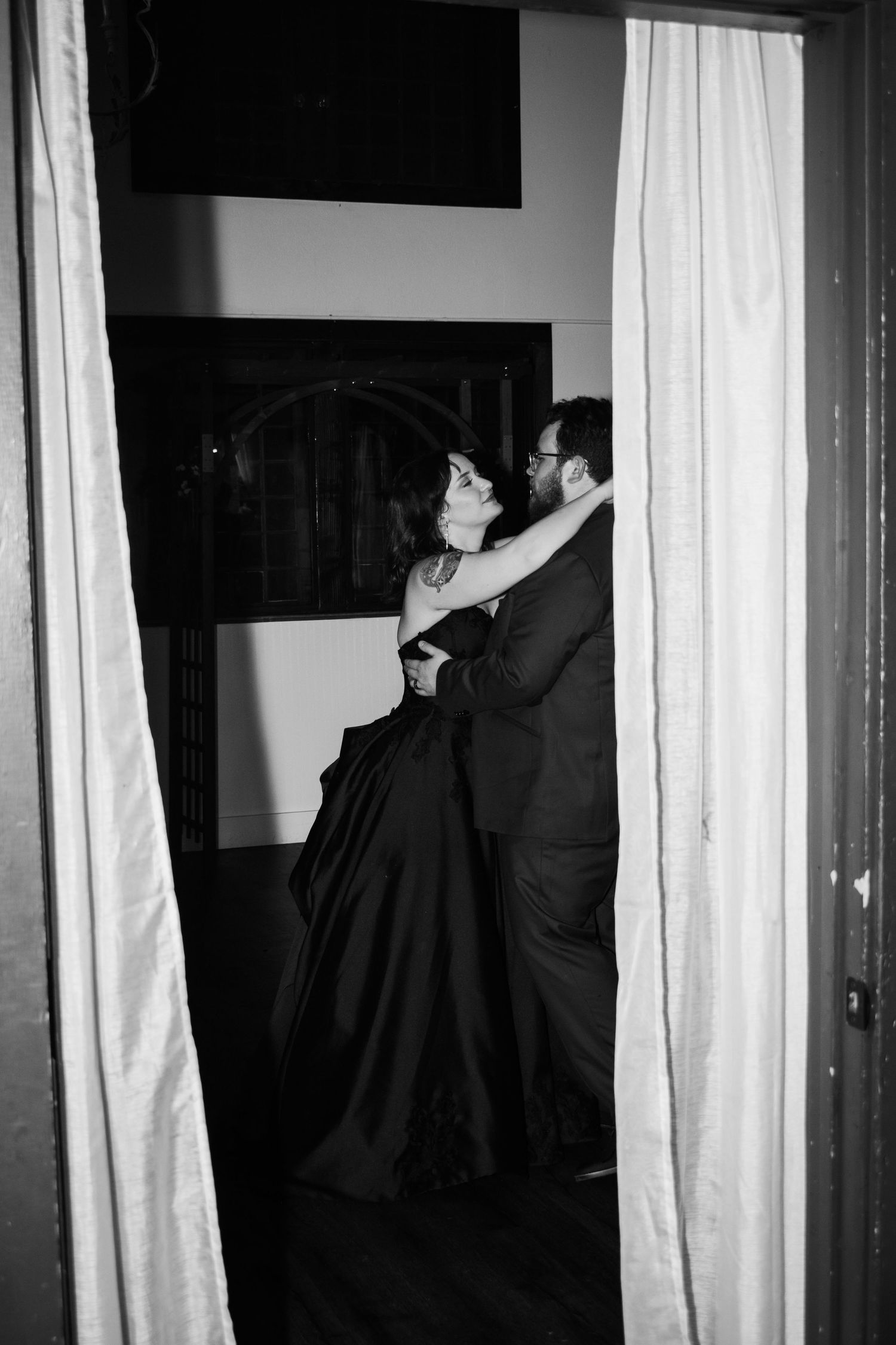 Black and white photo of an elegant couple embracing in a doorway, woman in a long dark gown, man in a suit.