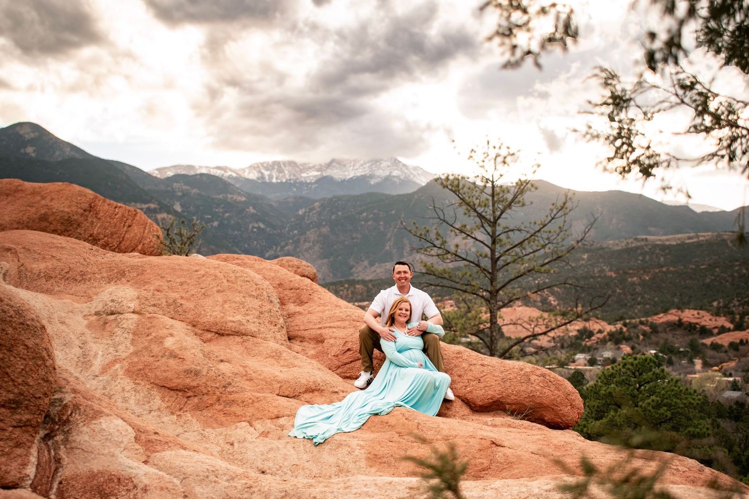 Couple poses for maternity session on red rocks at Garden of the Gods with Pikes Peak and dramatic cloudy sky backdrop.