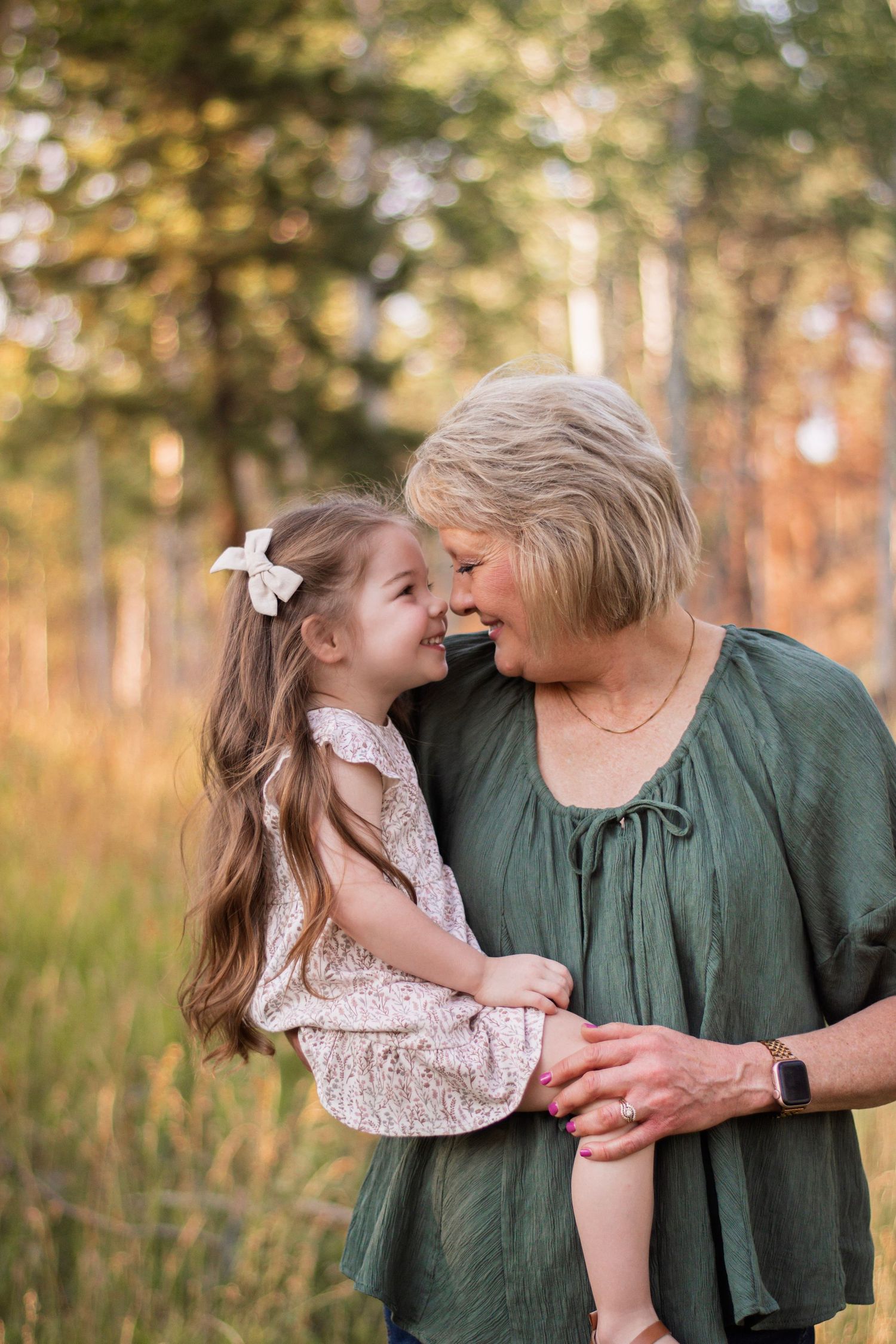 Grandmother holding young girl with long hair and bow, touching noses and smiling in a sunlit forest meadow.