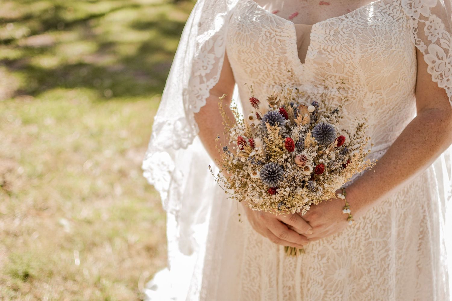 Bride in lace wedding dress holding a rustic dried flower bouquet with thistles and wildflowers outdoors.