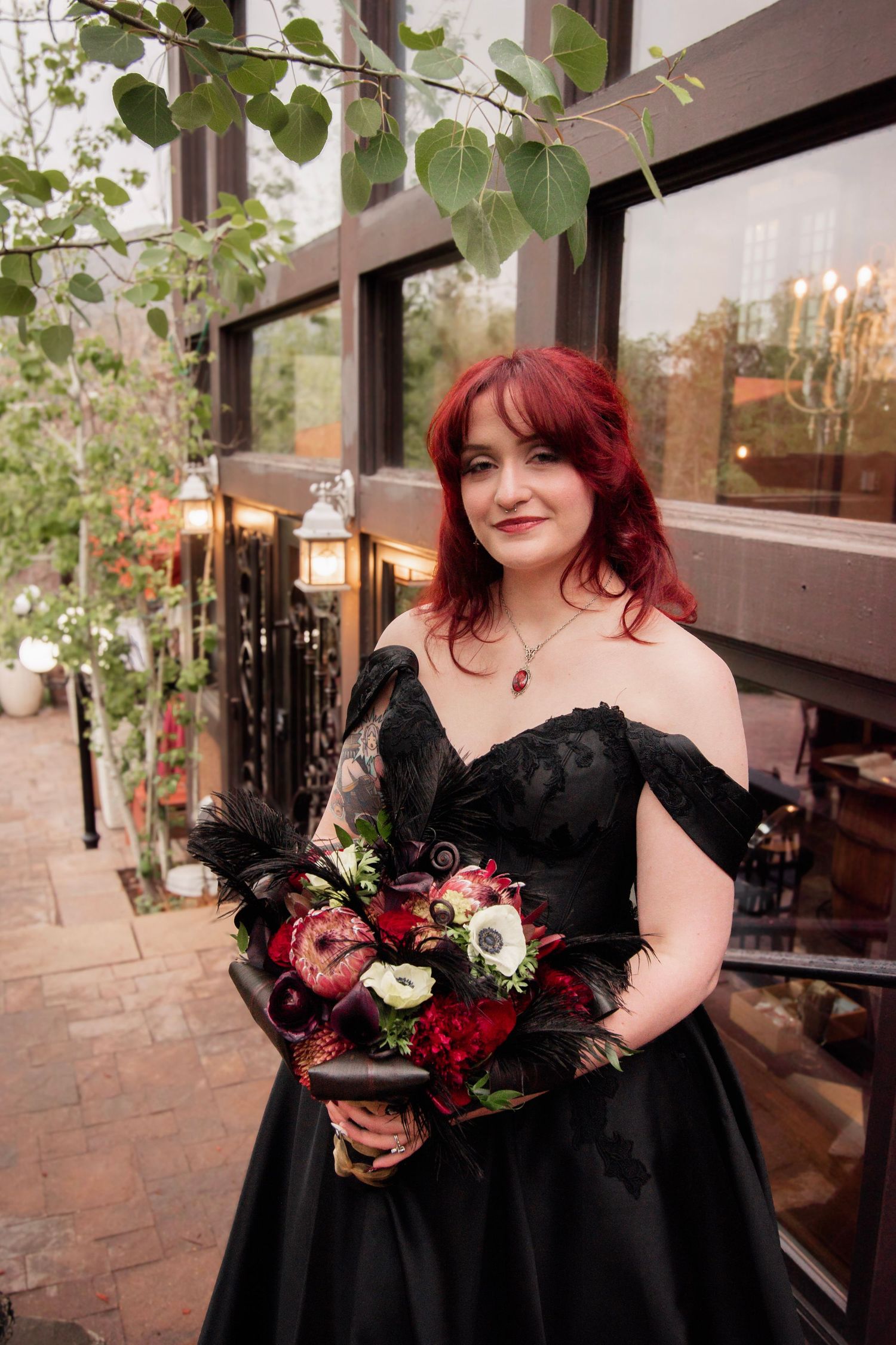 A woman with red hair in a black off-shoulder gown holds a dark floral bouquet outdoors at an elegant venue.