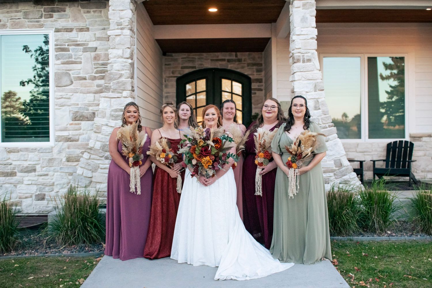 Bride in white gown poses with five bridesmaids in mismatched jewel-tone dresses holding autumn bouquets.