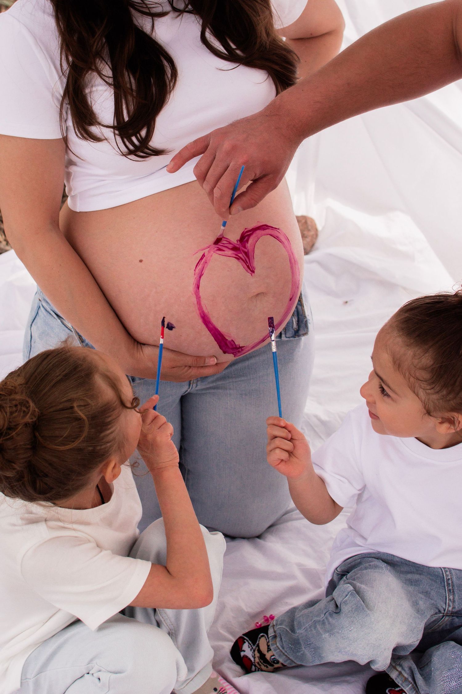 Two children painting a pink heart on their pregnant mother's belly with brushes.