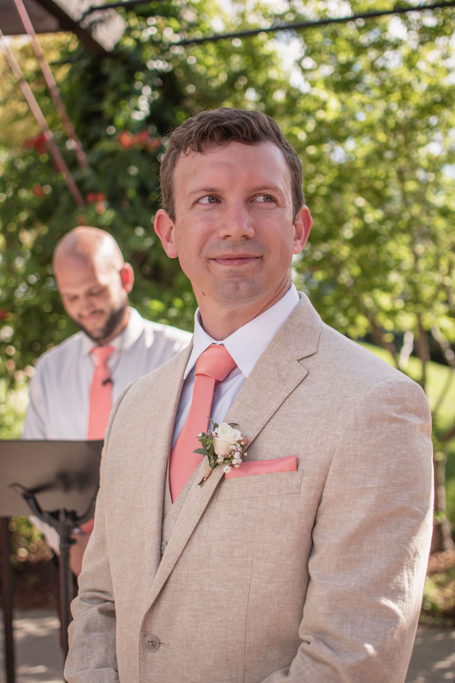 Handsome groom in tan suit with coral tie and boutonniere watching bride walk down aisle.
