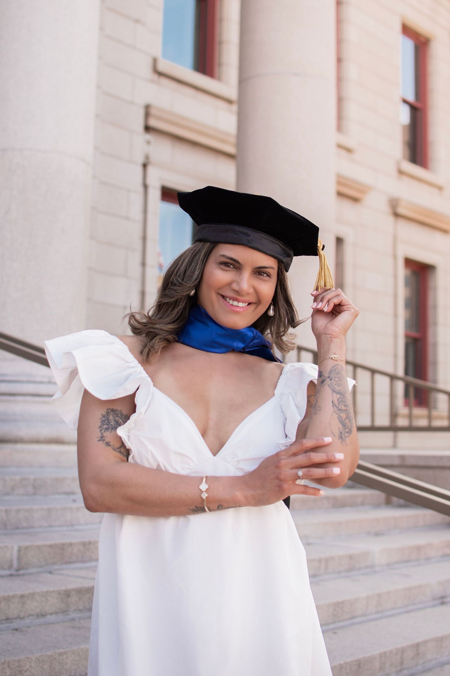 Smiling woman in white dress and black graduation cap holding tassel on university steps.
