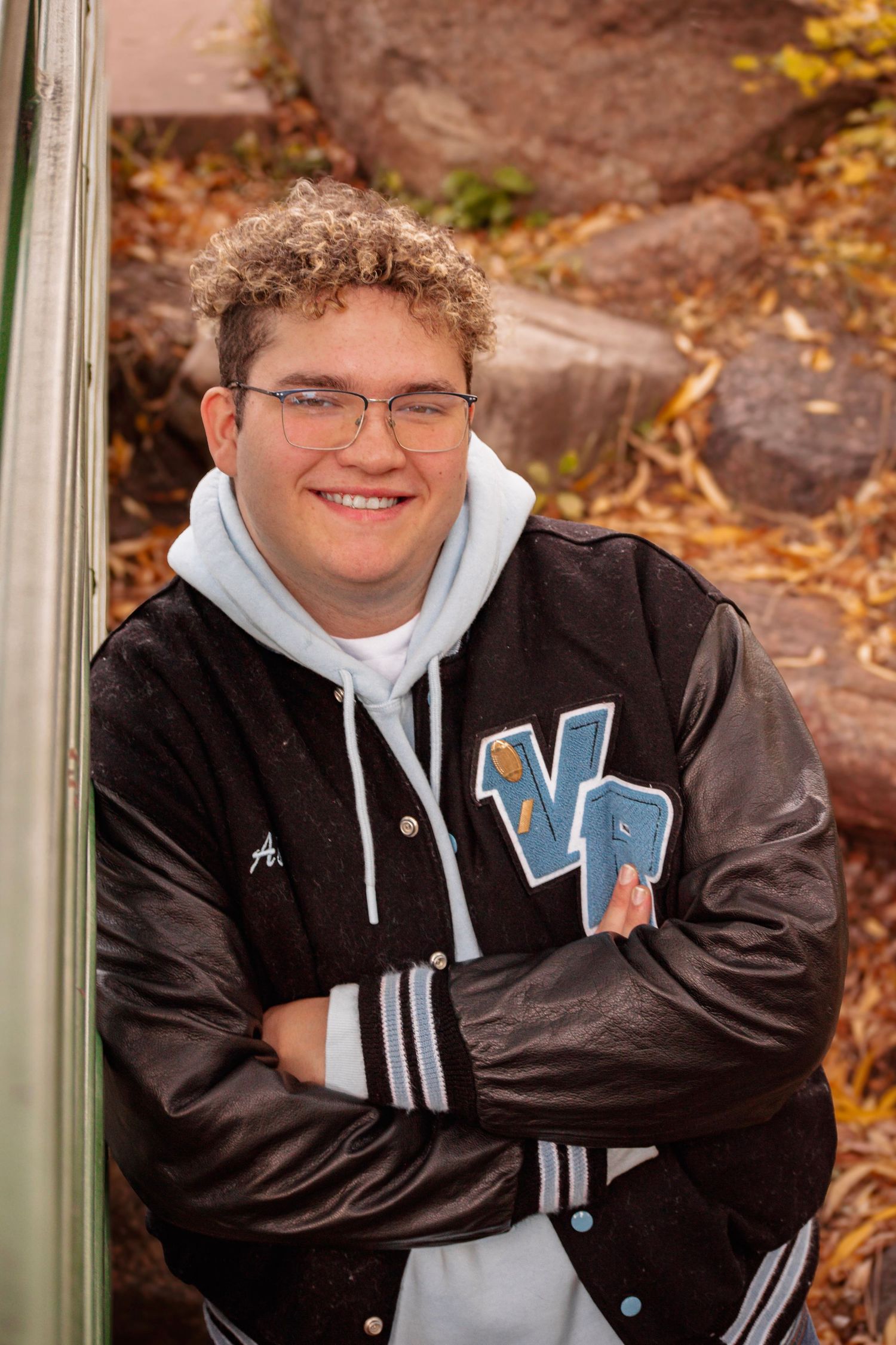 Smiling teen in glasses and varsity jacket with arms crossed, posing outdoors among autumn leaves and rocks.