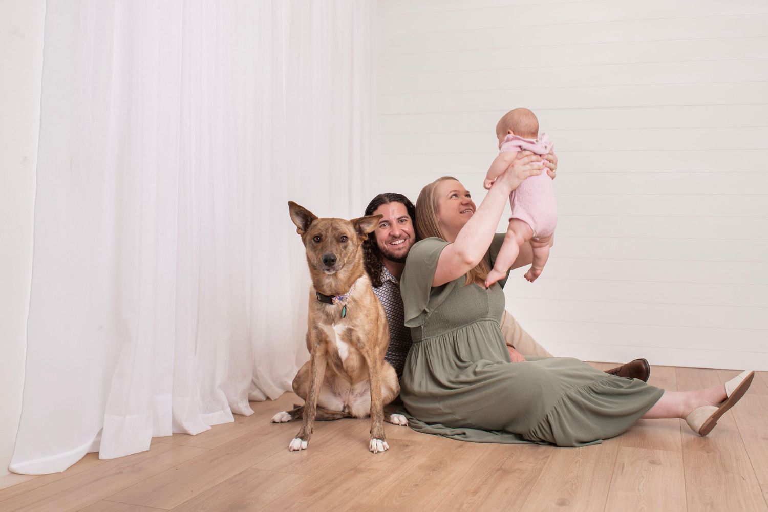 Two women sit on hardwood floor with a brown dog and hold a baby in pink outfit against white curtain backdrop at Luminous Creative Studios.