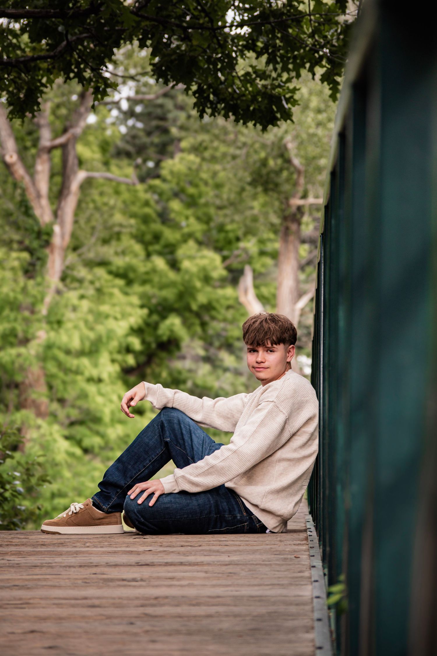 Young man in beige sweater and jeans sitting on wooden deck against green wall surrounded by lush trees.
