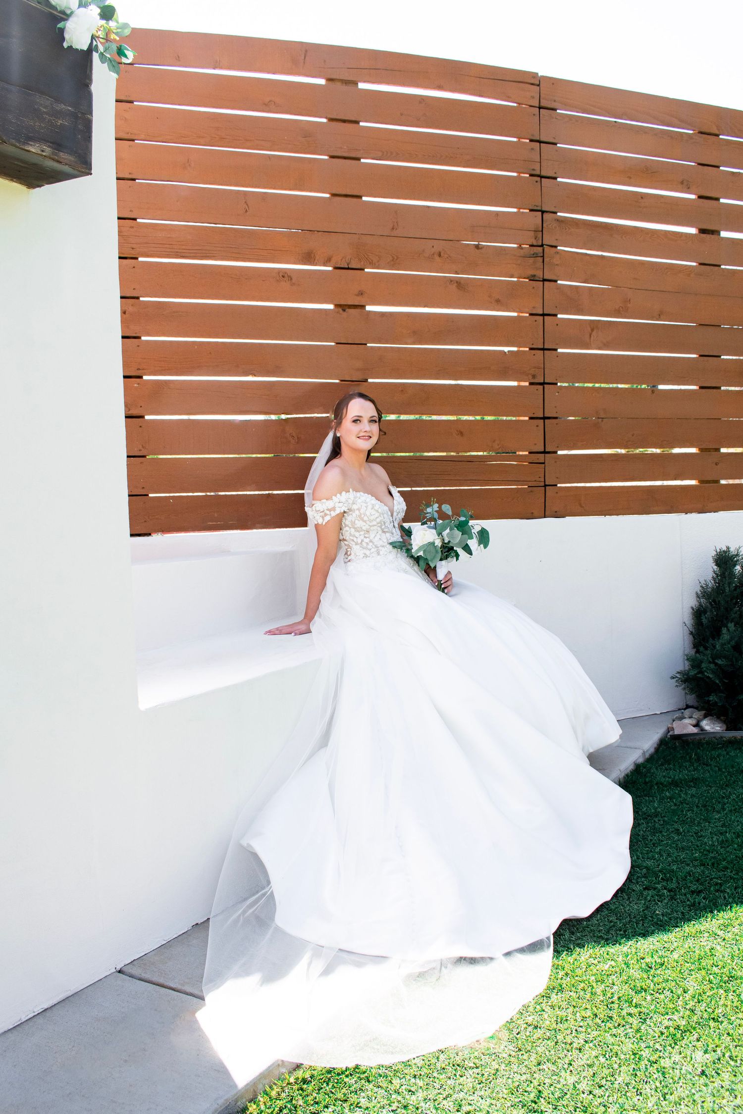 Bride in white off-shoulder ball gown holding bouquet, seated by wooden fence backdrop outdoors.