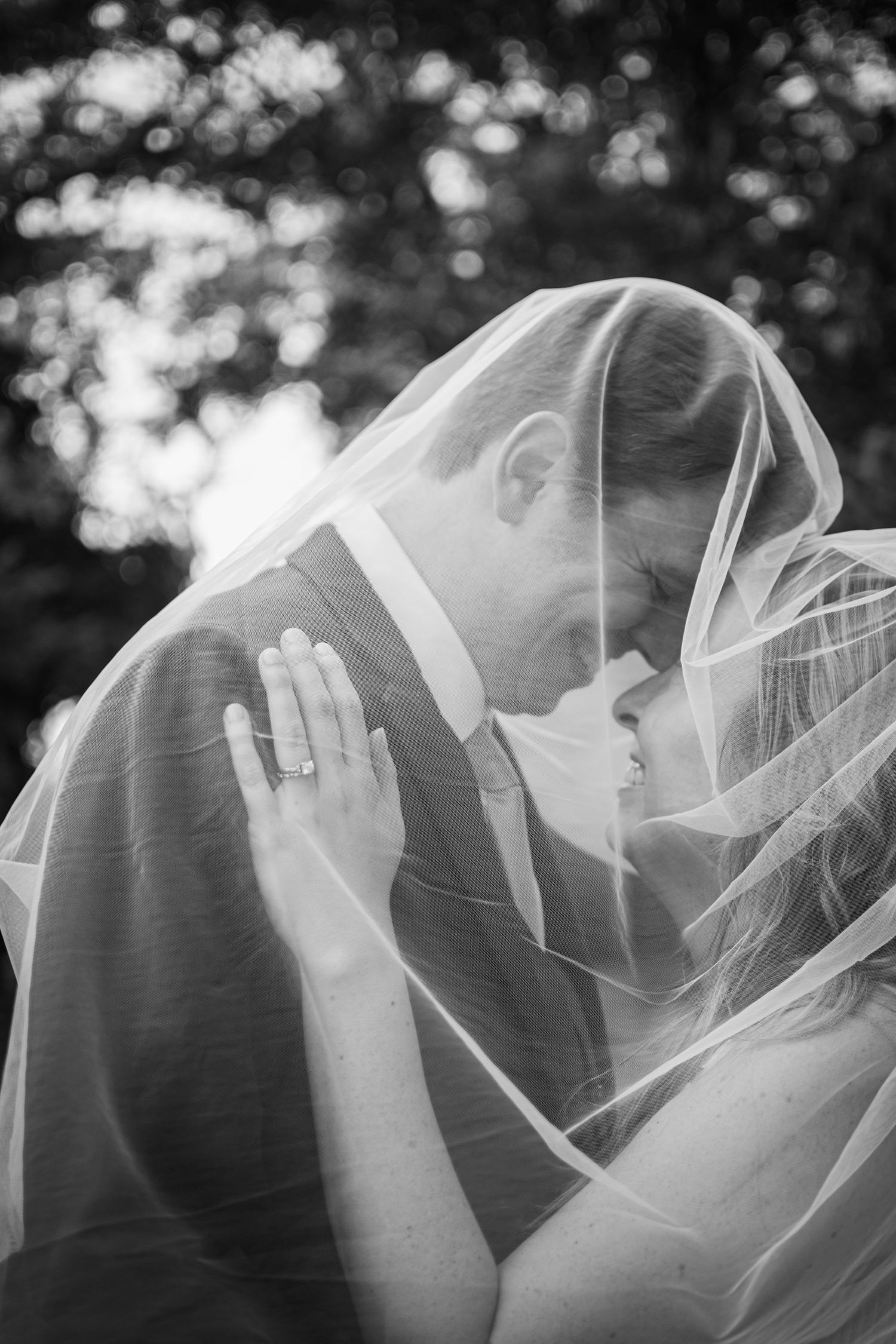 Black and white photo of bride and groom sharing intimate moment under wedding veil outdoors.