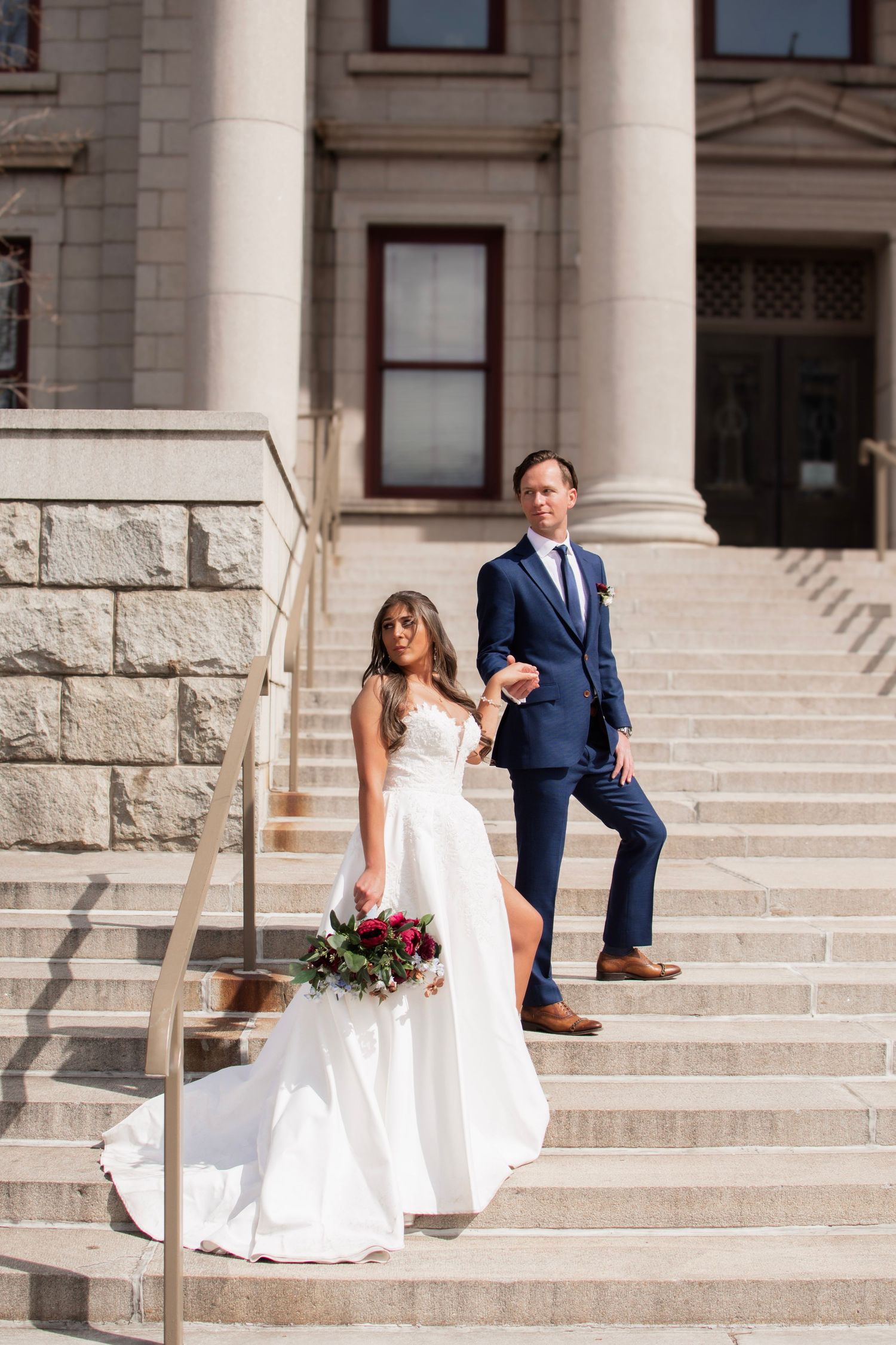 Bride and groom holding hands on stone steps of elegant building, bride in white gown holding red bouquet.