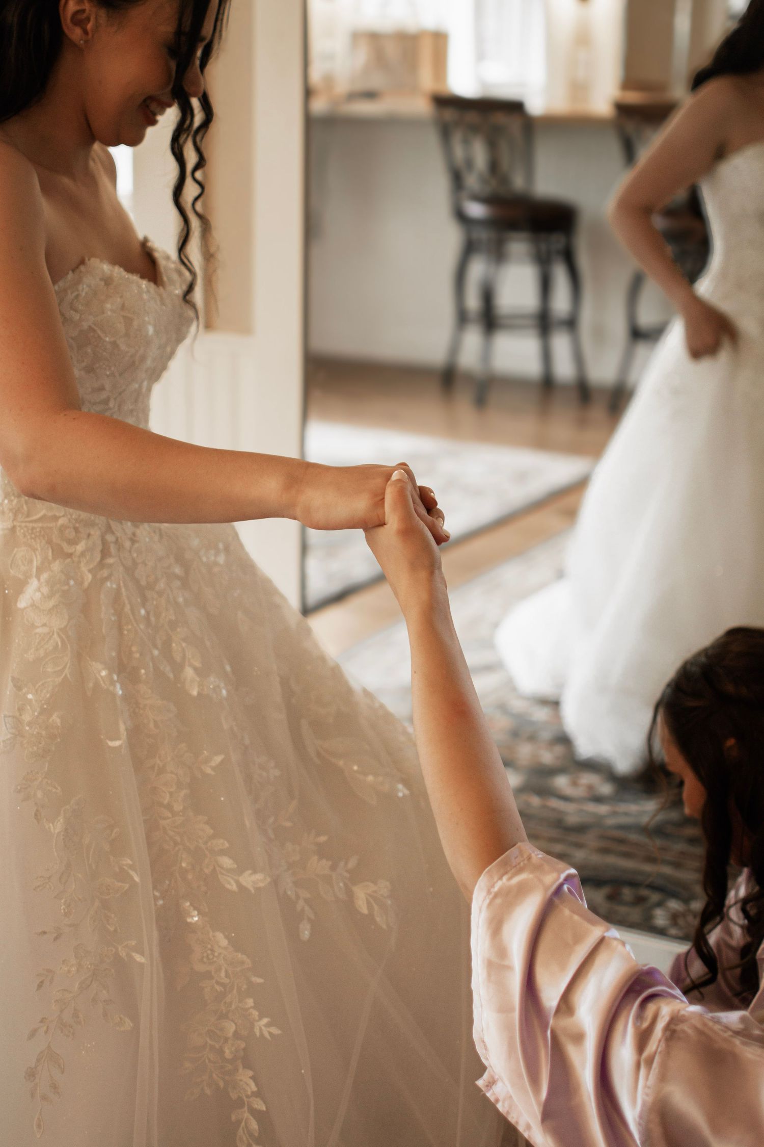 Bride in lace wedding gown holding hands with a young flower girl in a pink robe indoors.