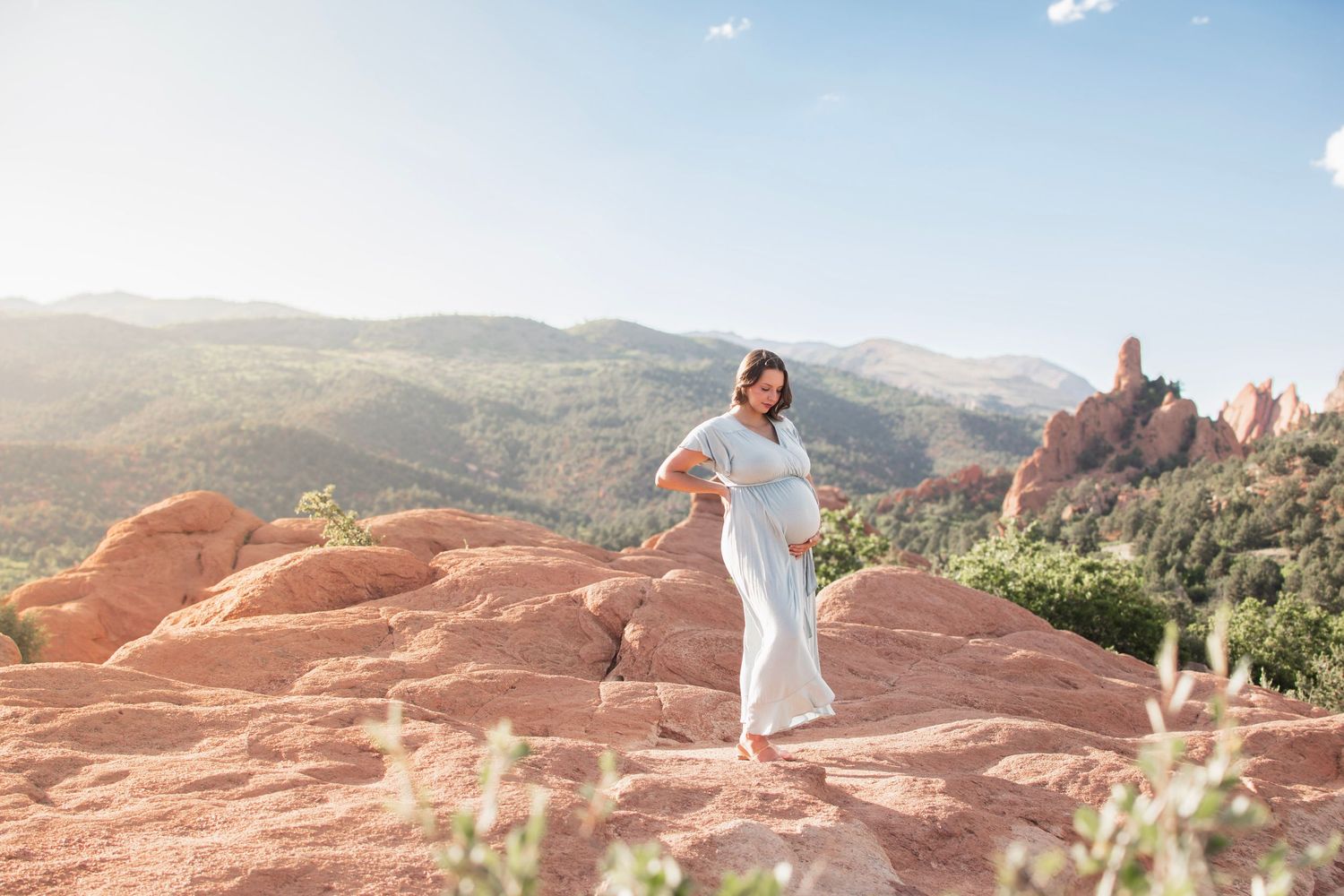 Pregnant woman in white dress poses on red rocks at Garden of the Gods, Colorado, with mountain views.