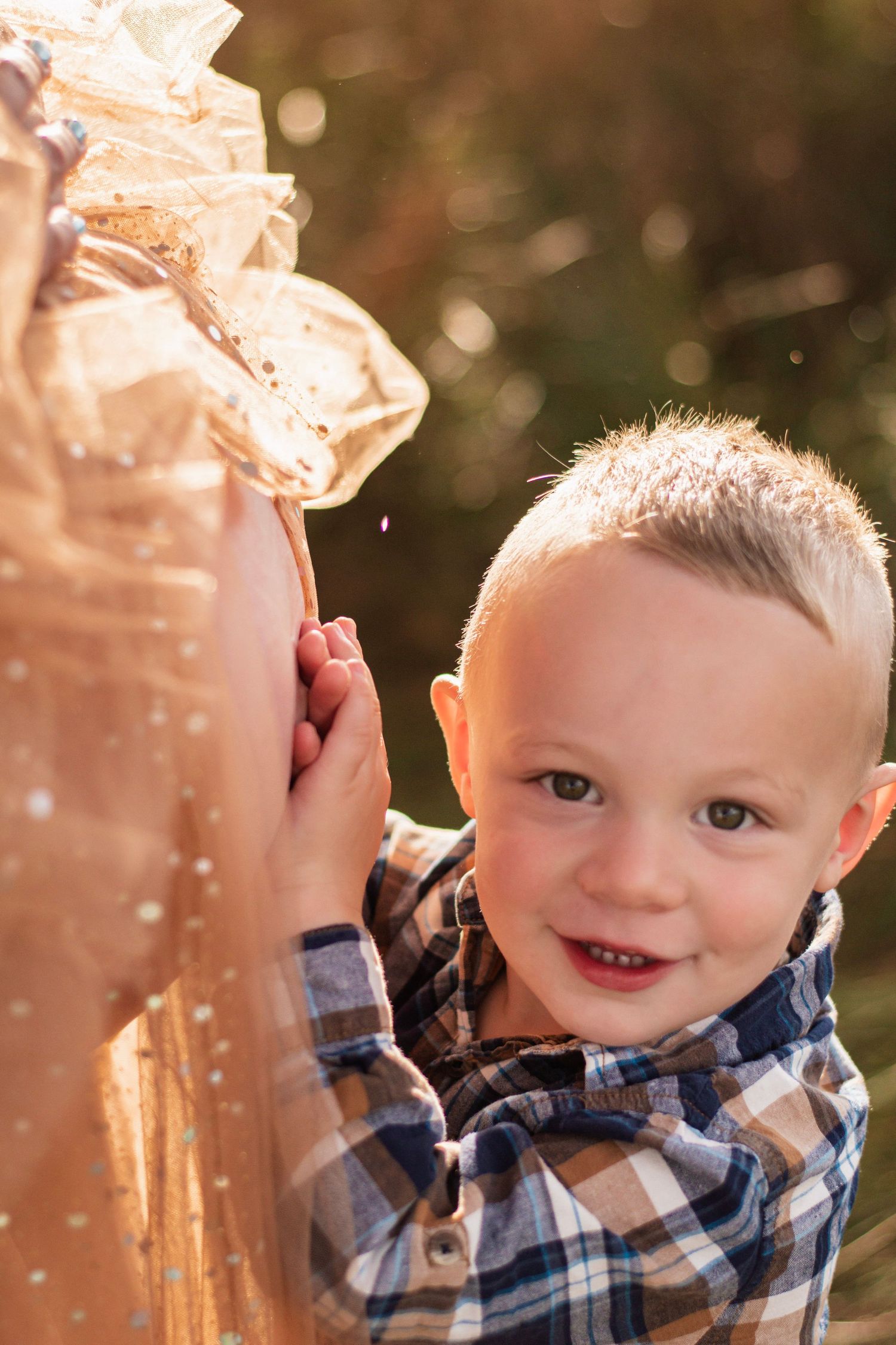 A smiling toddler boy in a plaid shirt holds his pregnant mothers belly outdoors in warm golden light.