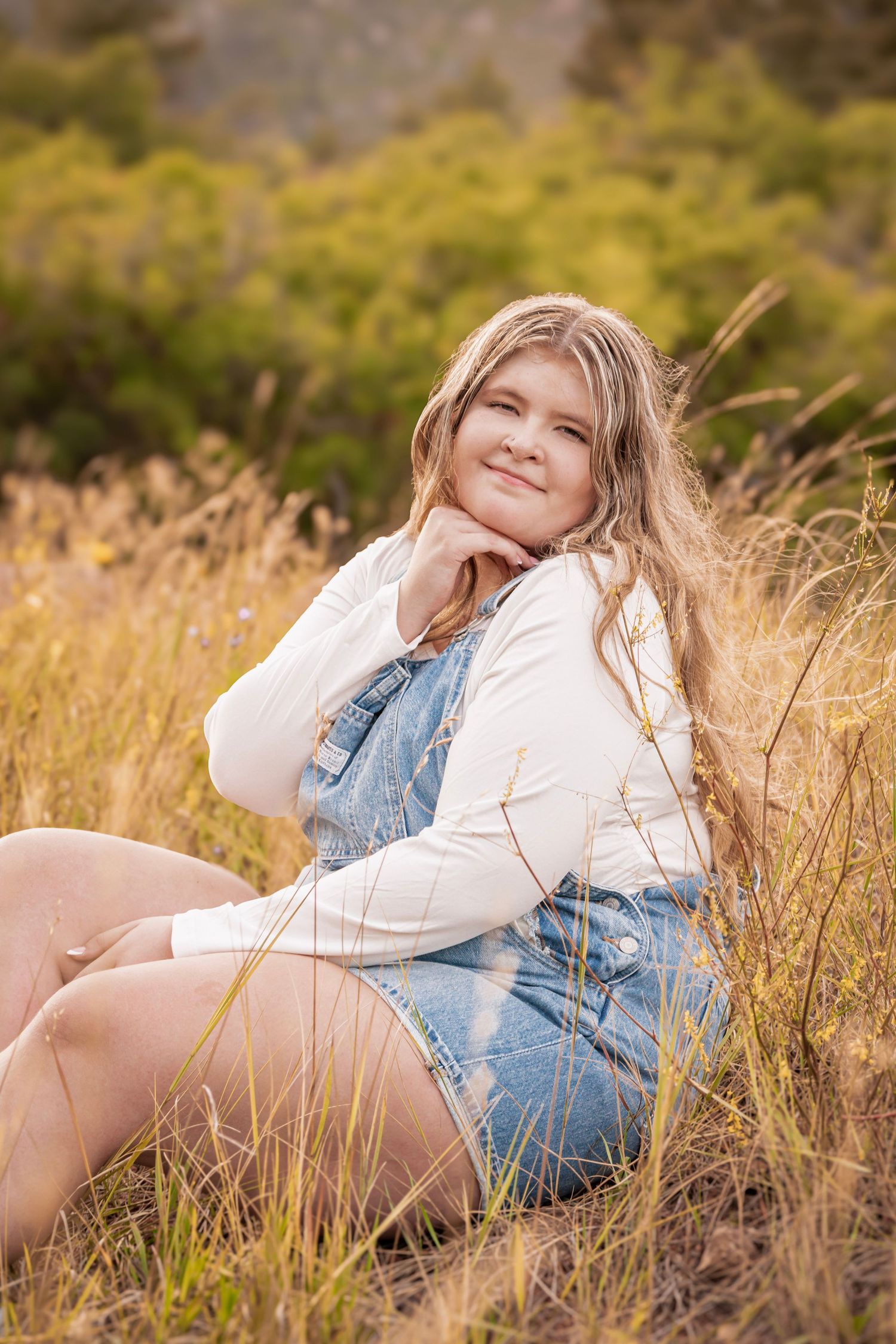 Young blonde girl in denim overalls sitting in golden grass field, smiling outdoors in natural setting at her senior photoshoot.