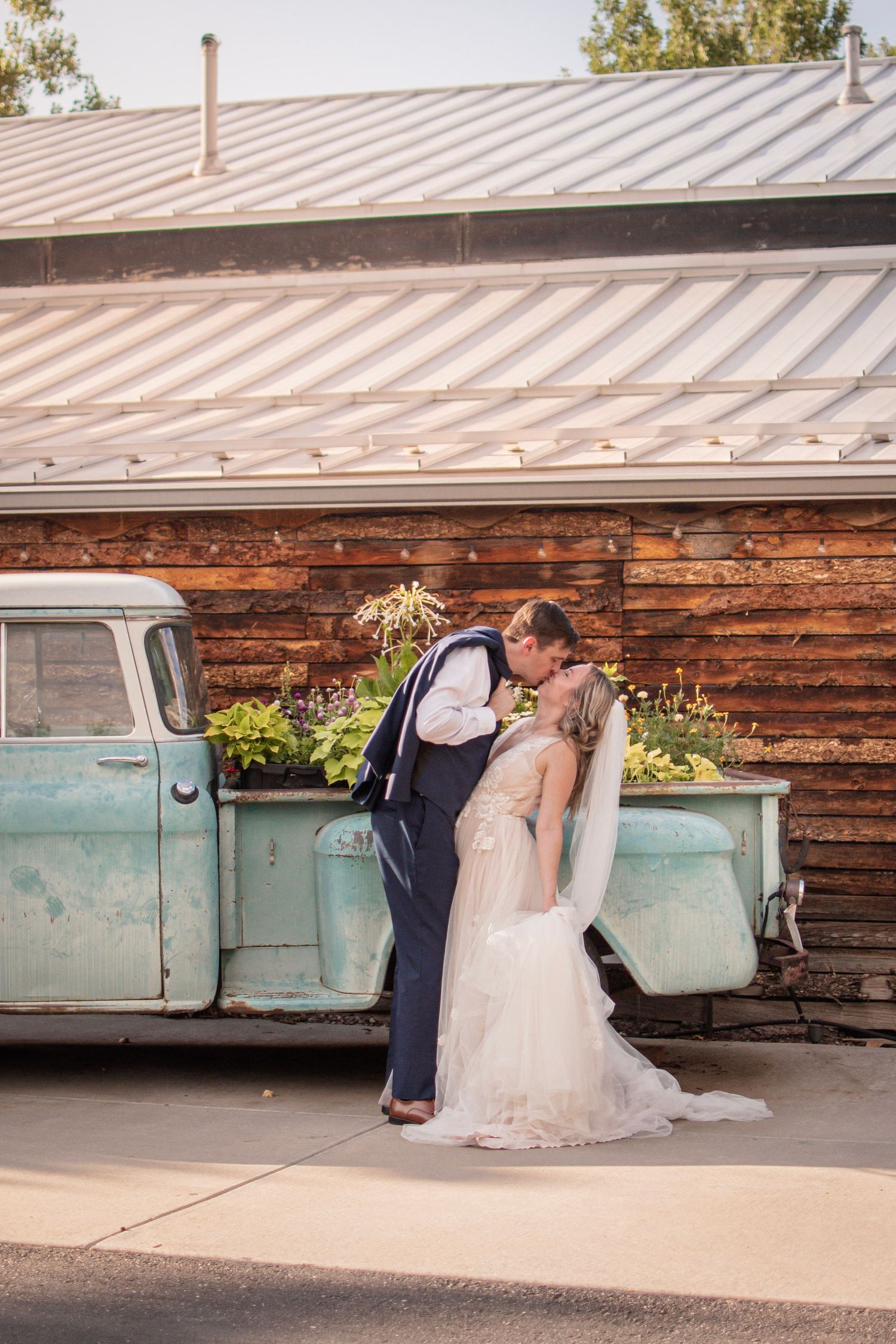 Bride and groom share a romantic kiss beside a vintage mint truck with flowers, in front of a rustic wooden barn.