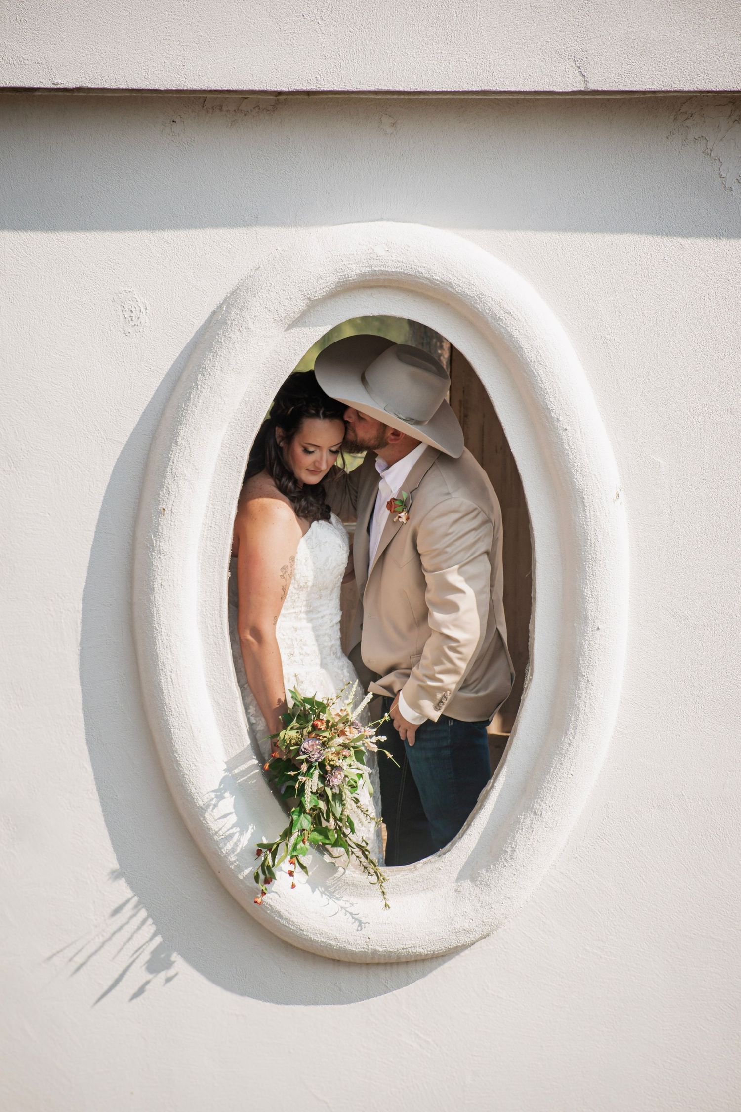 Bride and groom share a romantic moment framed by an oval window, with the groom in a cowboy hat holding a bouquet.