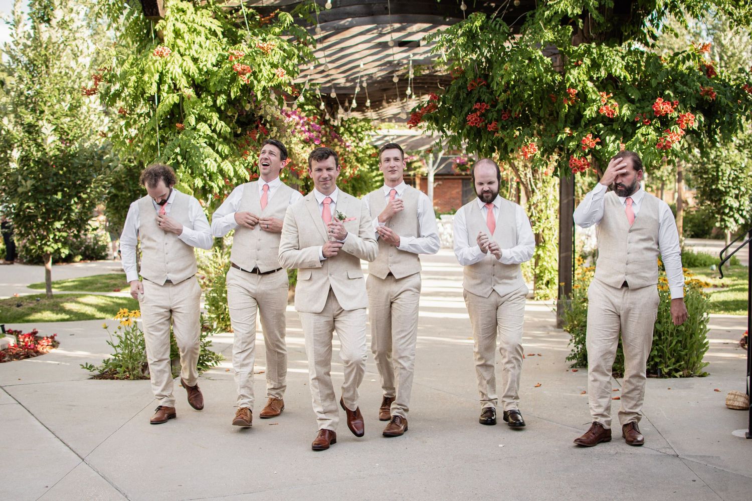 Six groomsmen in tan suits and pink ties walk together outdoors under a floral pergola at a wedding.
