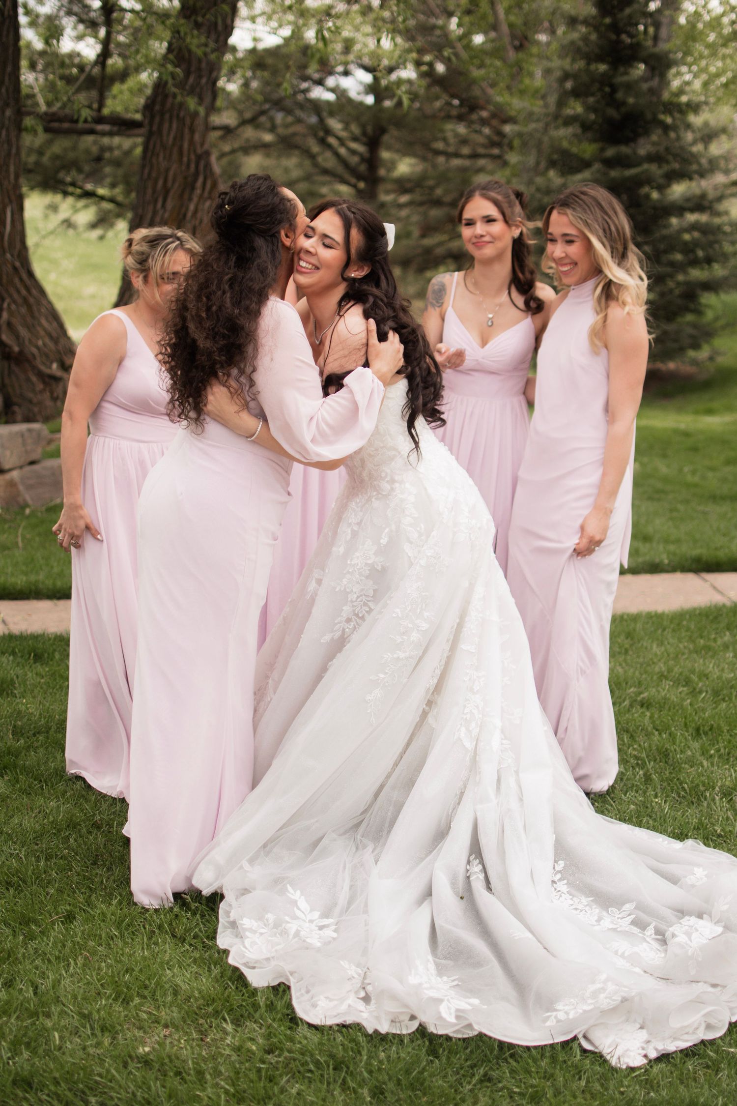 Bride in lace gown embracing bridesmaids in blush pink dresses outdoors on green grass.