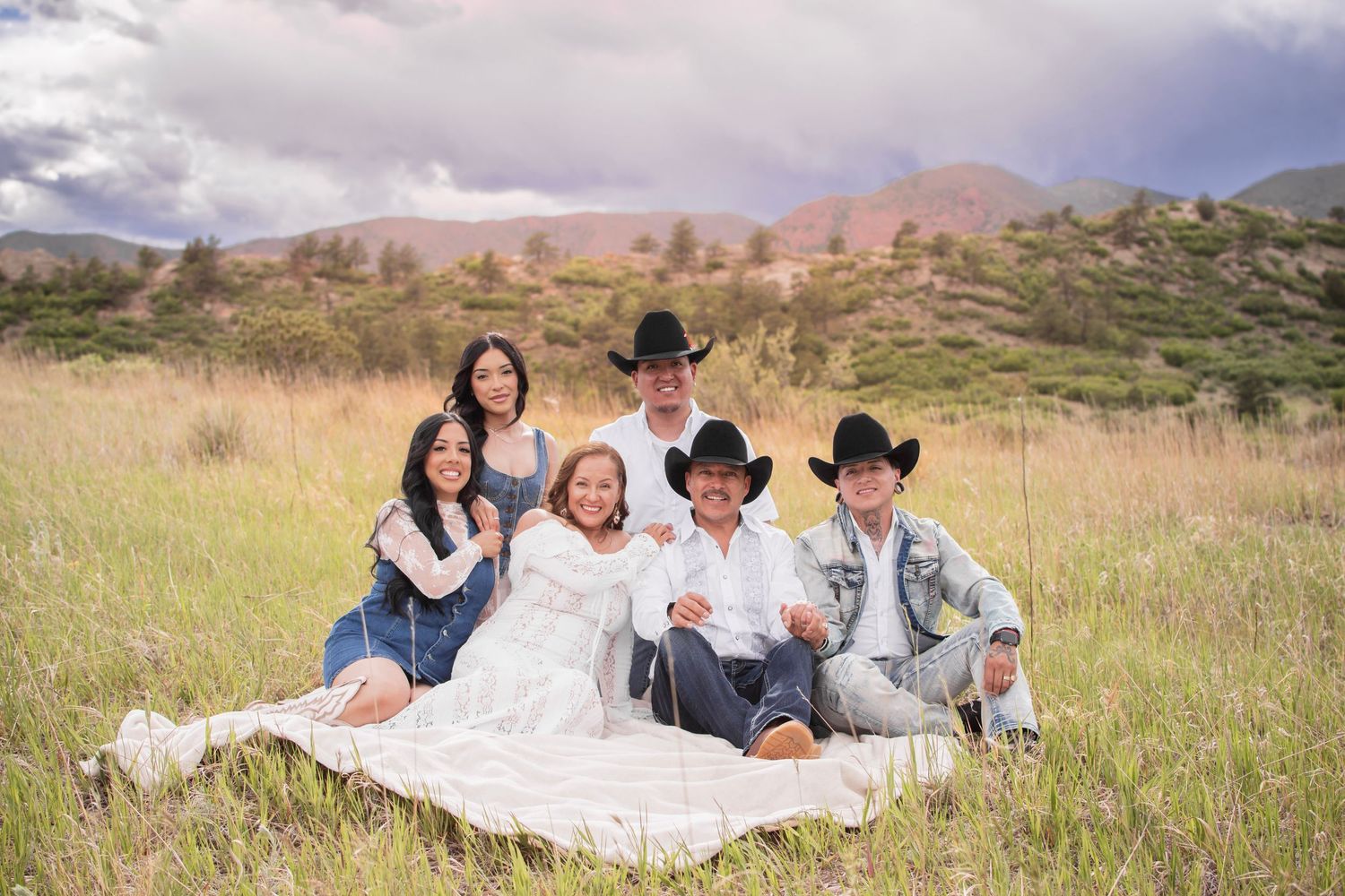 Family of six posing in a grassy mountain meadow, some wearing cowboy hats, sitting on a white blanket outdoors at Ute Valley Park.