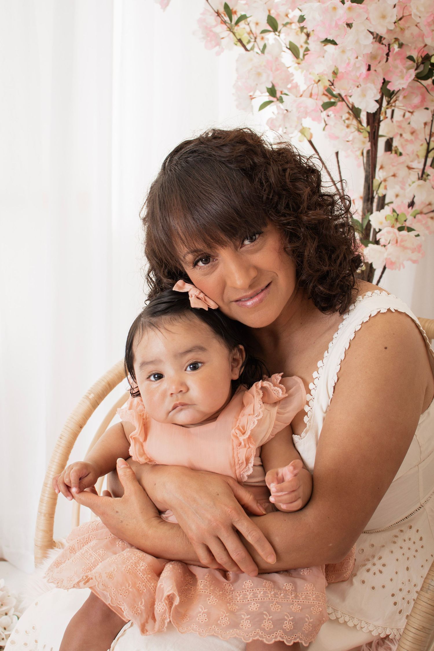 A smiling woman with curly hair holds a baby girl in a pink dress, posing before cherry blossom branches.