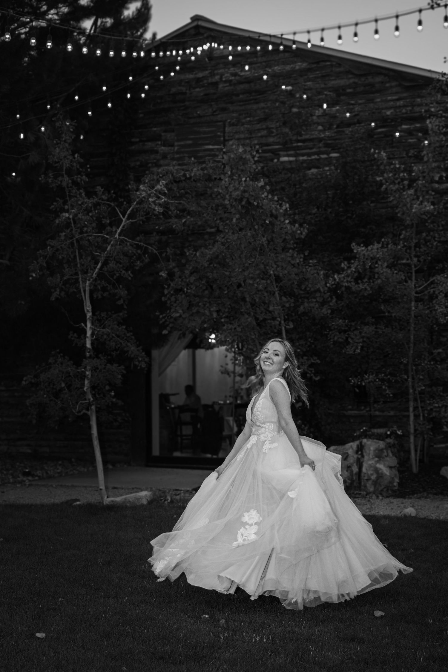 Bride twirls in a flowing white gown at dusk near a rustic barn with string lights above.