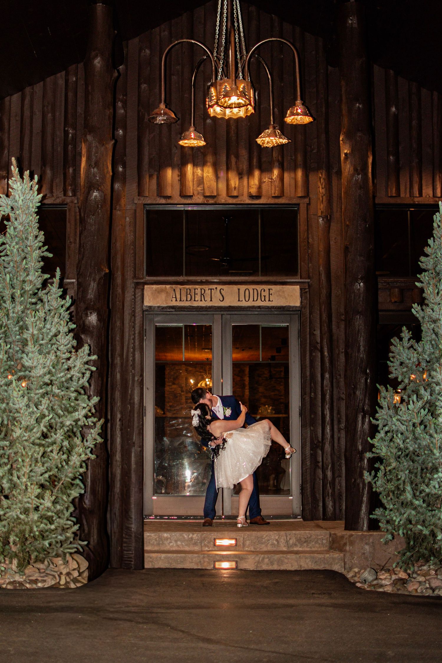 Couple sharing a romantic dip kiss at night outside Albert's Lodge rustic wooden entrance with warm pendant lights.
