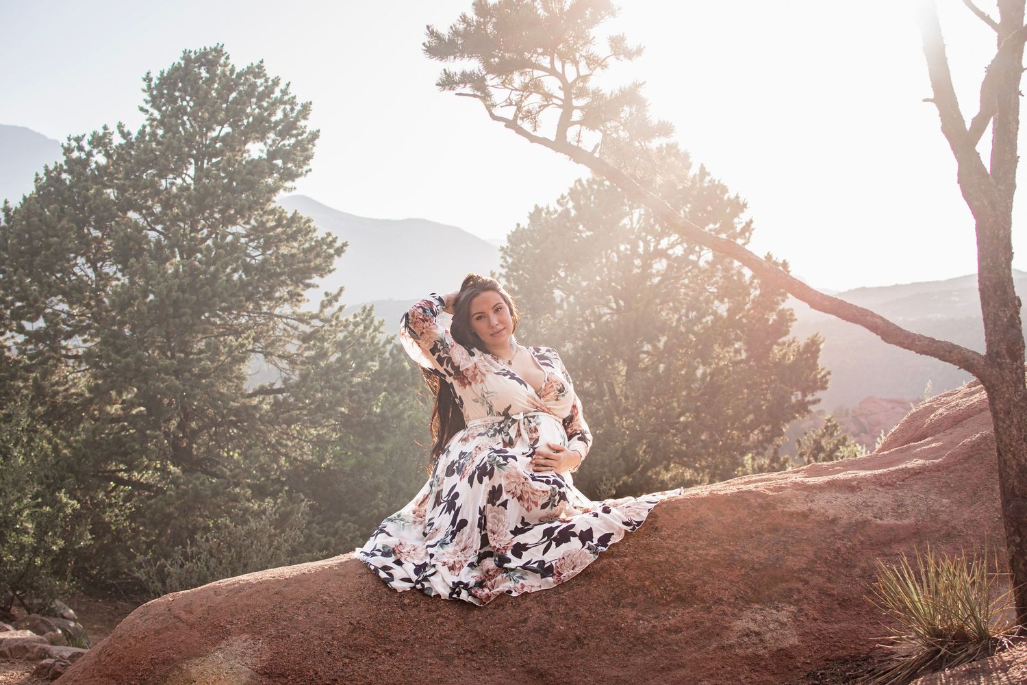 Woman in a flowing floral dress sits on a red sandstone rock among pine trees and mountains in warm golden sunlight.