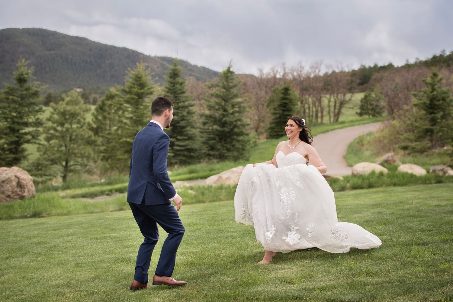 Bride in white gown and groom in navy suit share a joyful moment on green lawn with mountain backdrop.