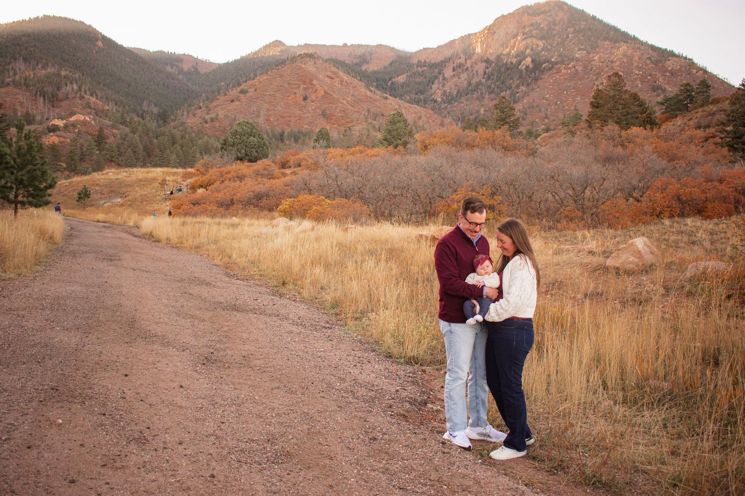 Family of three poses on a dirt path with golden fields and Colorado mountain backdrop at sunset at Blodgett Open Space