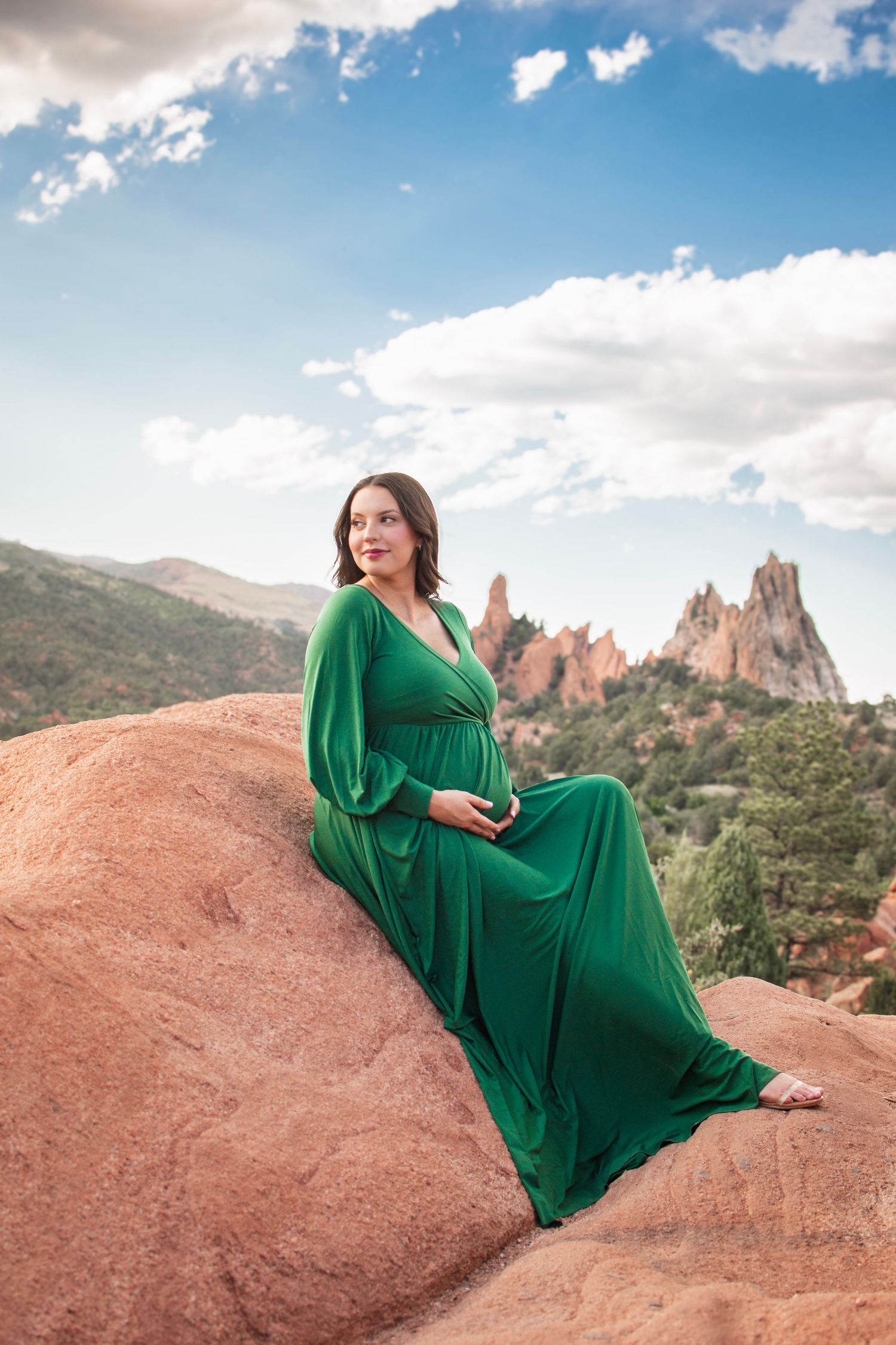 Pregnant woman in green dress sitting on red rocks at Garden of the Gods, Colorado, with dramatic rock formations behind her.