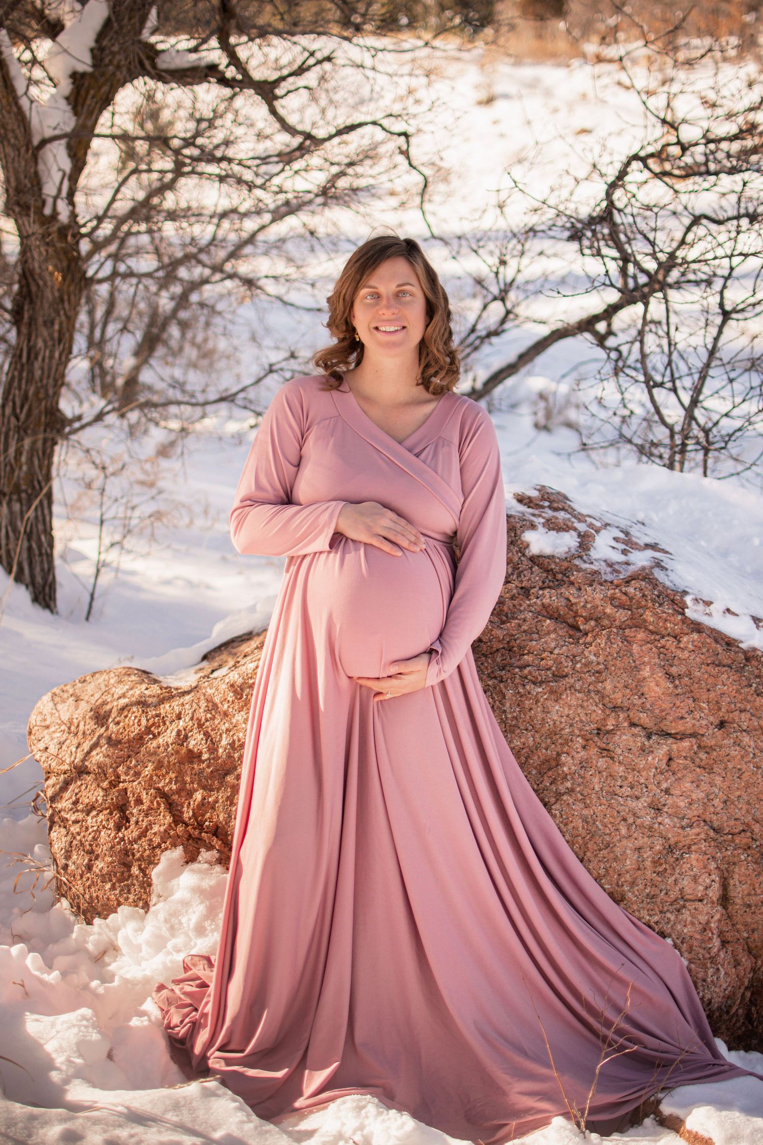 Pregnant woman in a flowing dusty rose maternity gown cradling her belly beside a boulder in a snowy winter landscape.