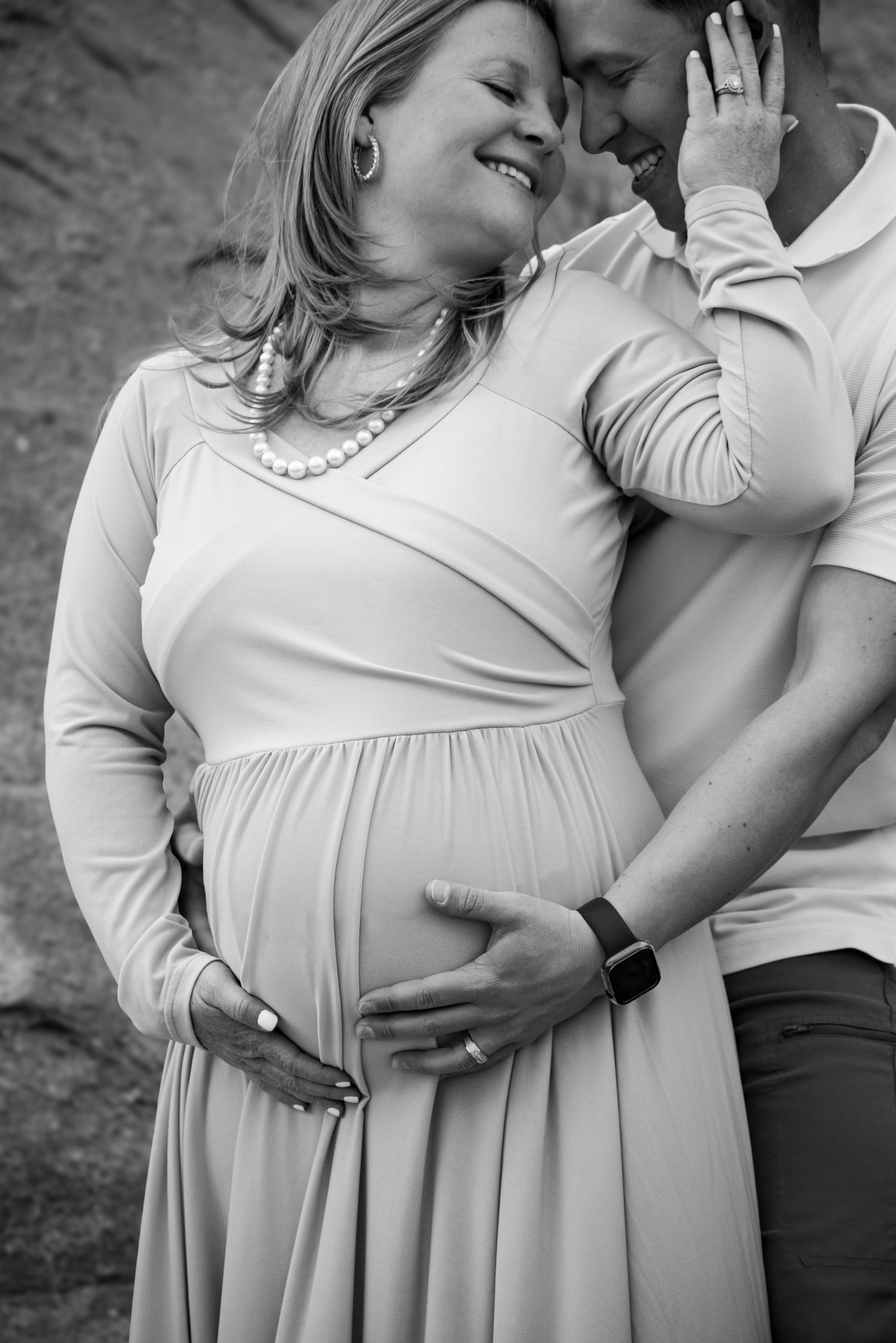 Black and white maternity photo of a smiling pregnant woman in a dress, embraced by her partner outdoors.
