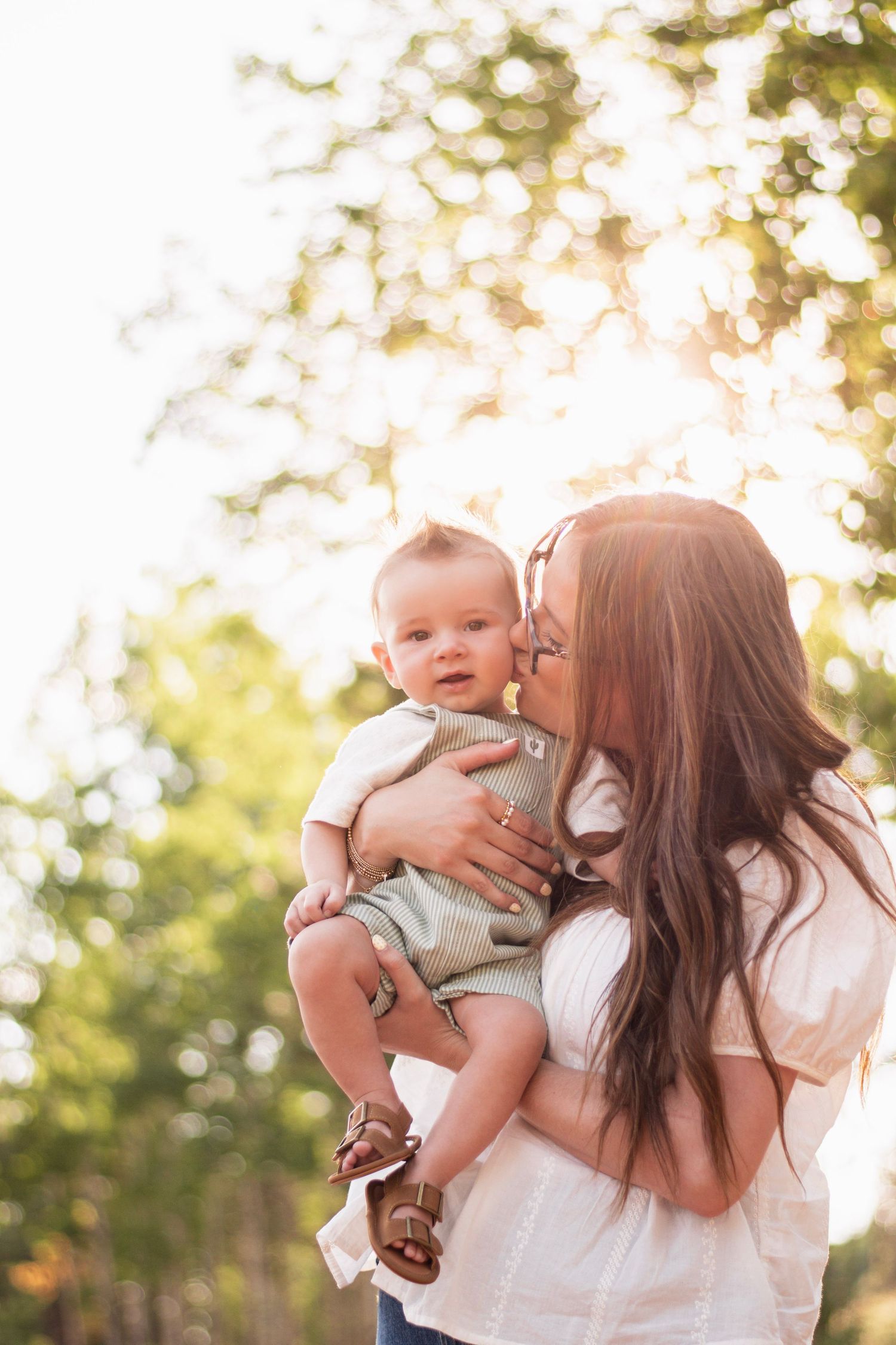 A mother tenderly kisses her baby outdoors in golden sunlight, surrounded by lush green trees.