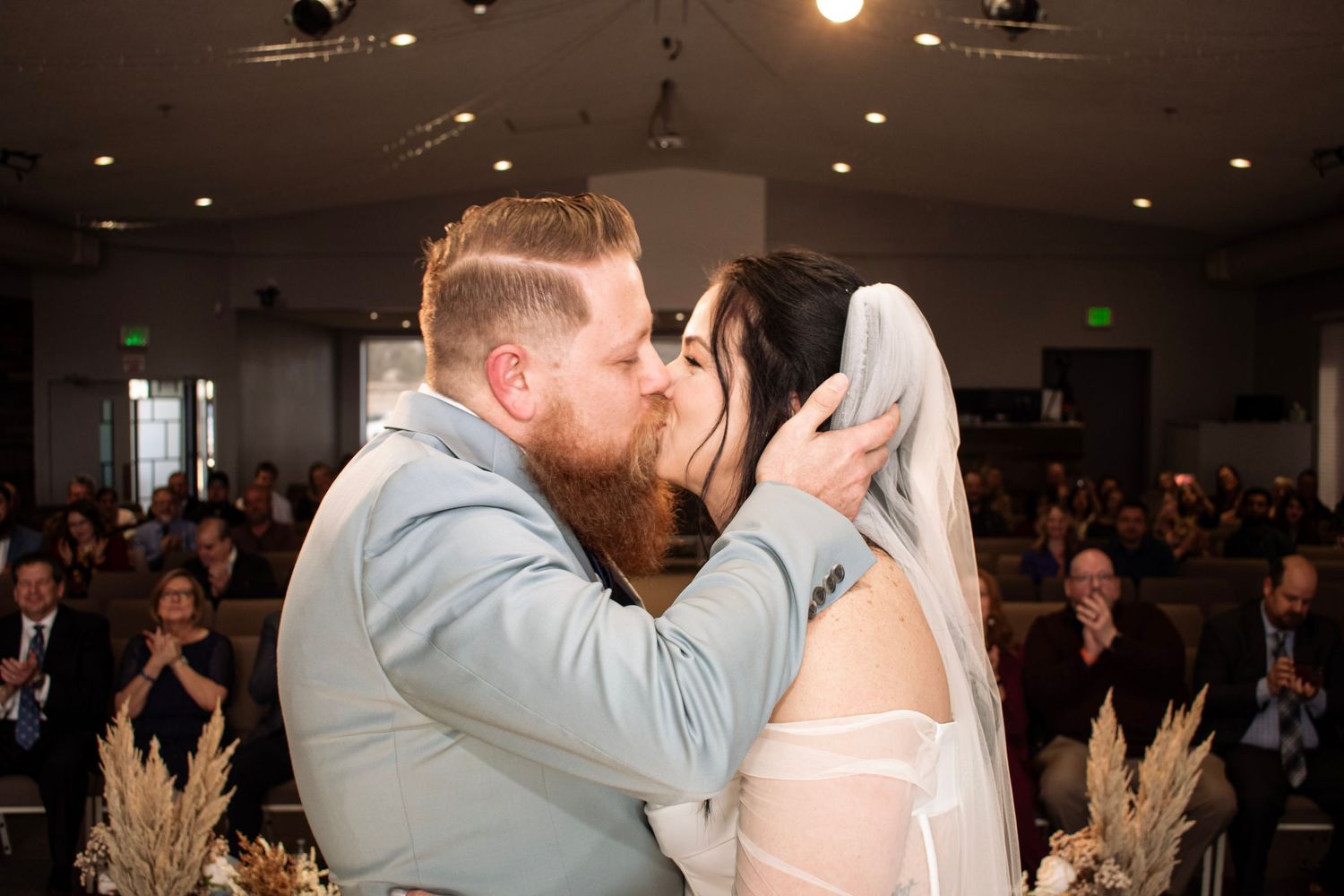 Bride and groom share first kiss at church wedding altar, guests applauding in background.